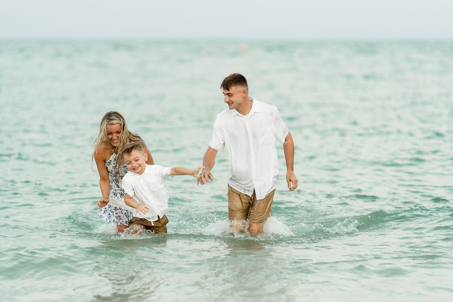 Family of three splashing and playing in ocean waves at the beach, parents and young child enjoying a sunny day.