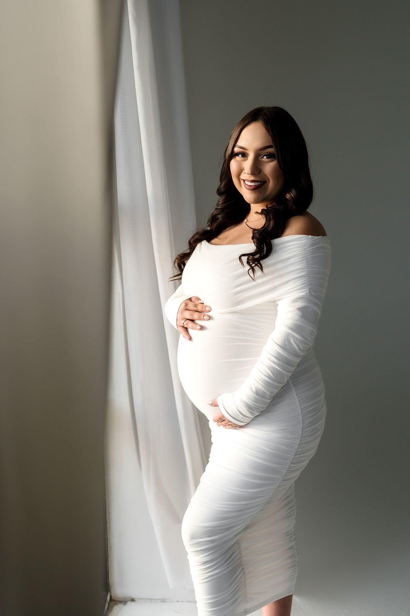 Smiling pregnant woman in white ruched off-shoulder dress cradling baby bump beside sheer curtains.