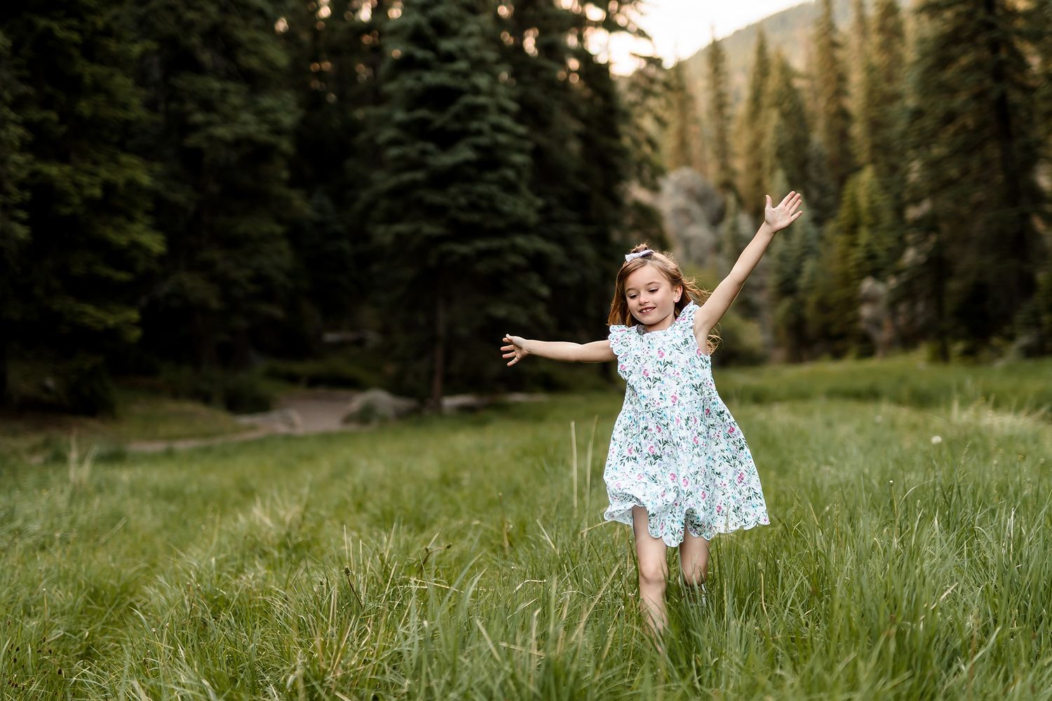 A joyful little girl in a floral dress runs through a lush green meadow with arms raised, surrounded by tall pine trees.