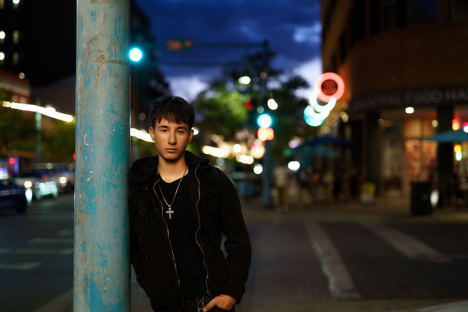 Teen boy in black jacket leaning against blue pole on city street at night with bokeh lights.