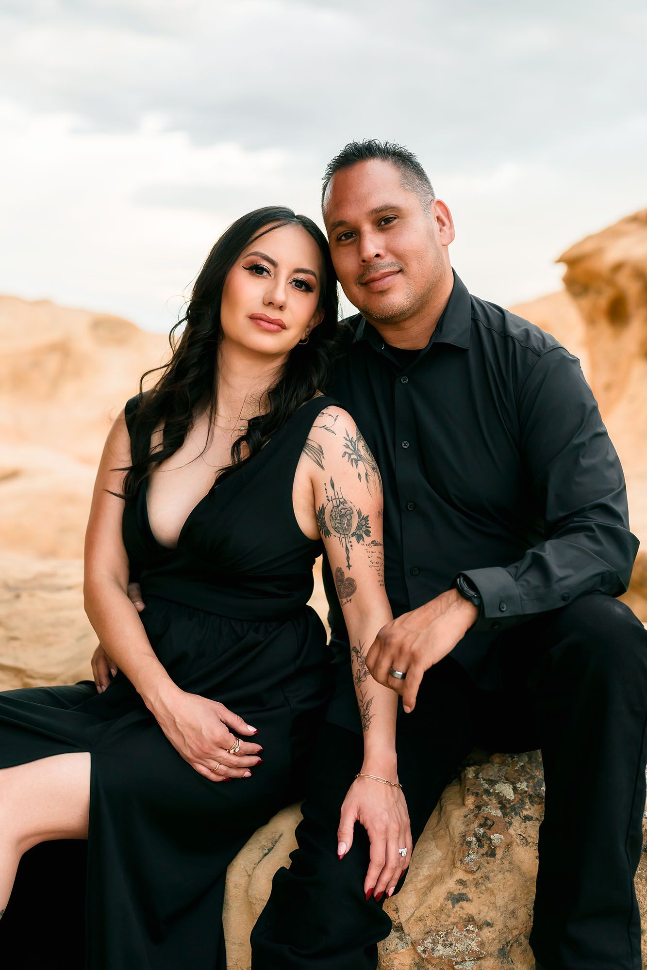Couple posing on rocks in matching black outfits, woman with arm tattoos leans against man, cloudy sky background.