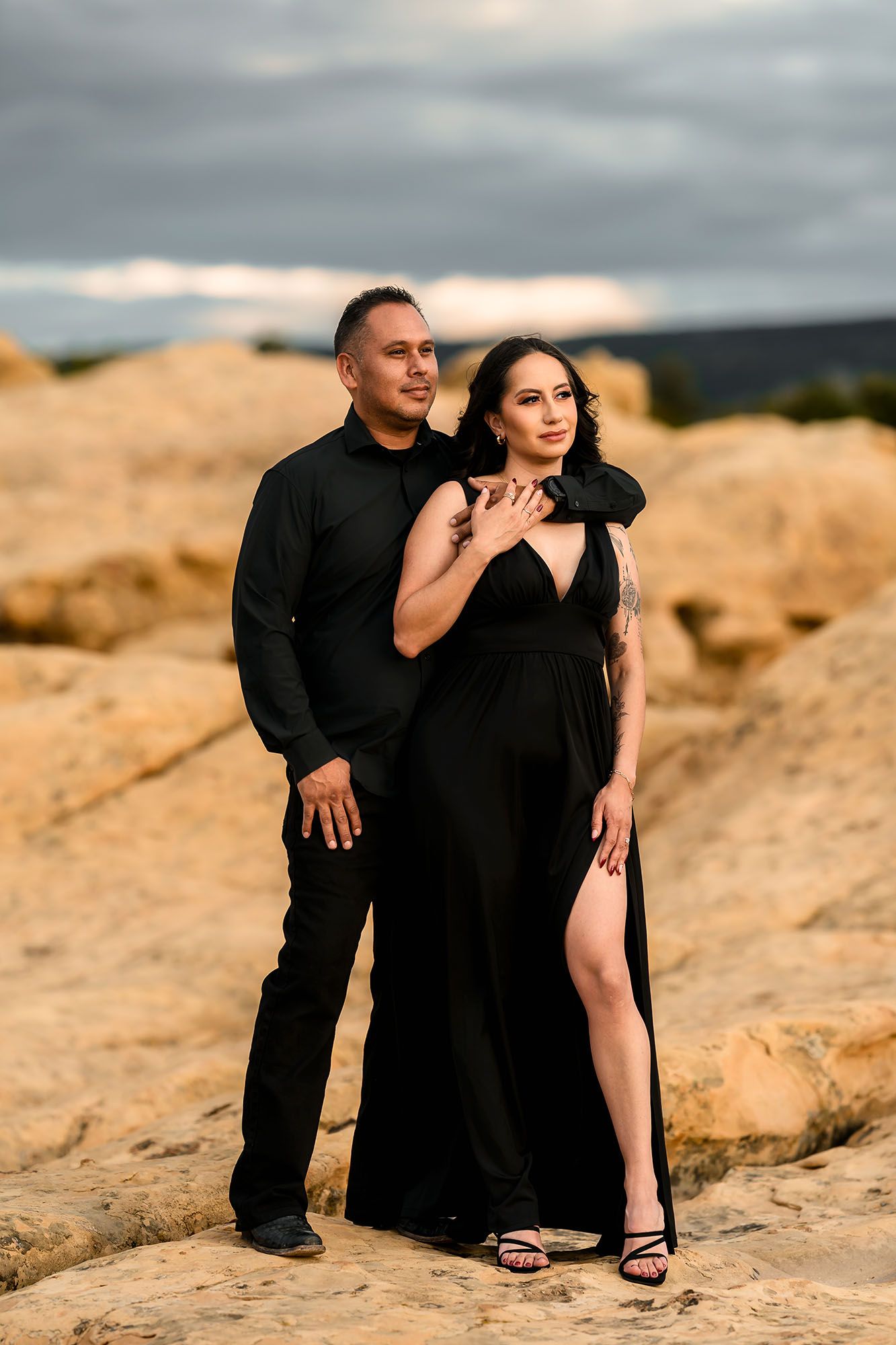 Couple in matching black outfits posing on rocky terrain under dramatic cloudy sky.
