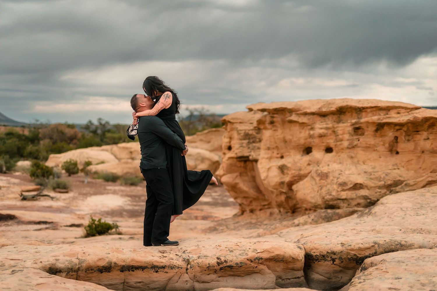 Man lifting woman in black dress on sandstone rocks with dramatic cloudy sky in desert landscape.