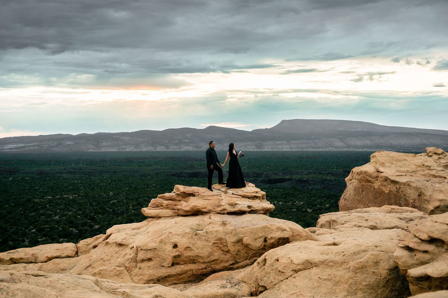 Couple in black standing on sandstone cliff overlooking vast green valley under cloudy sky.