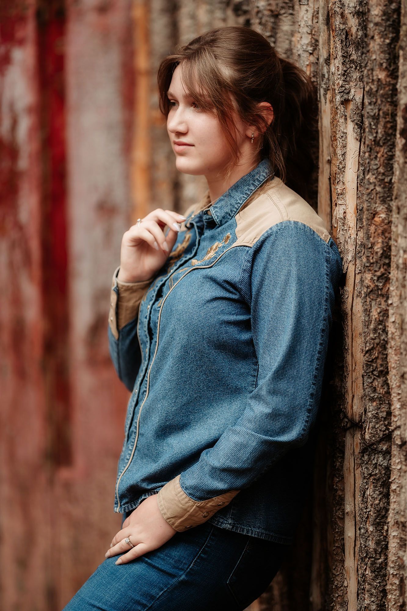 Brunette woman in denim jacket leaning against rustic wooden wall in moody portrait.