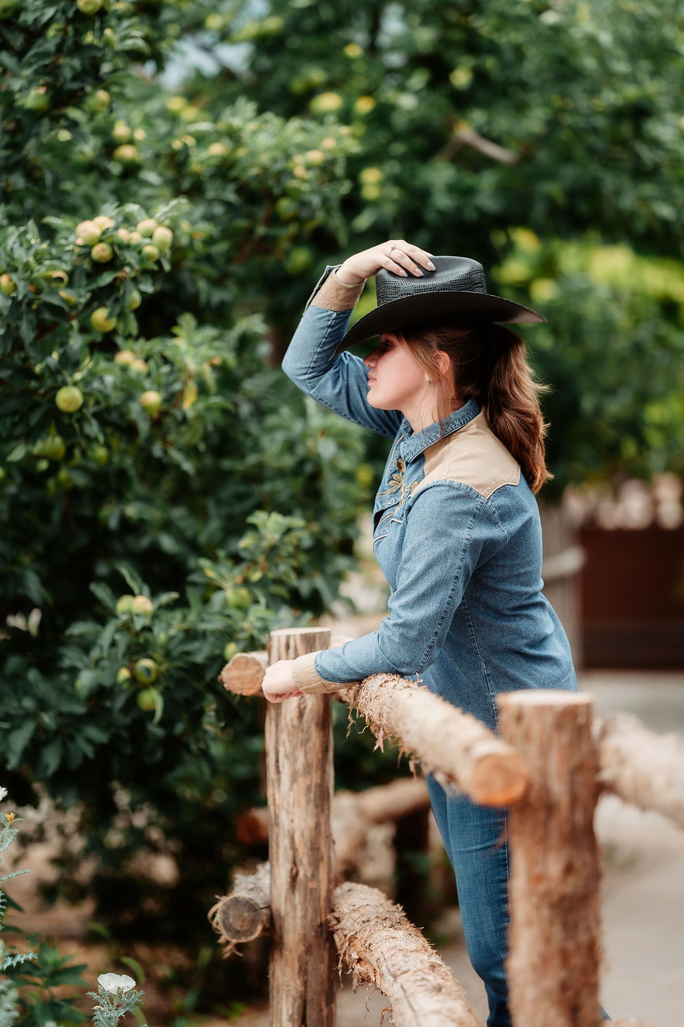 Woman in denim outfit and black cowboy hat leaning on wooden fence near apple trees.