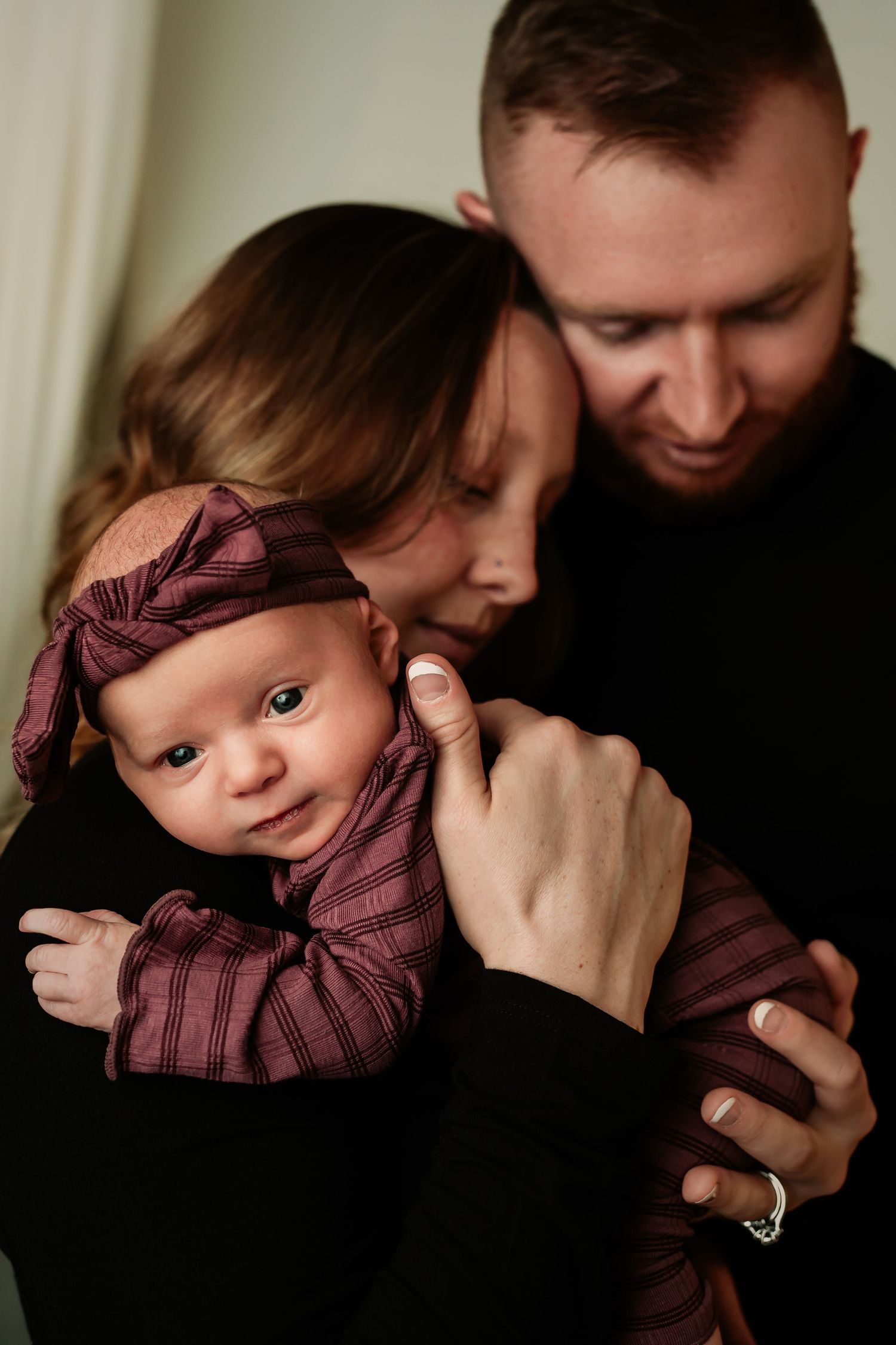 Intimate family portrait showing tender embrace with baby wearing burgundy outfit against dark background.