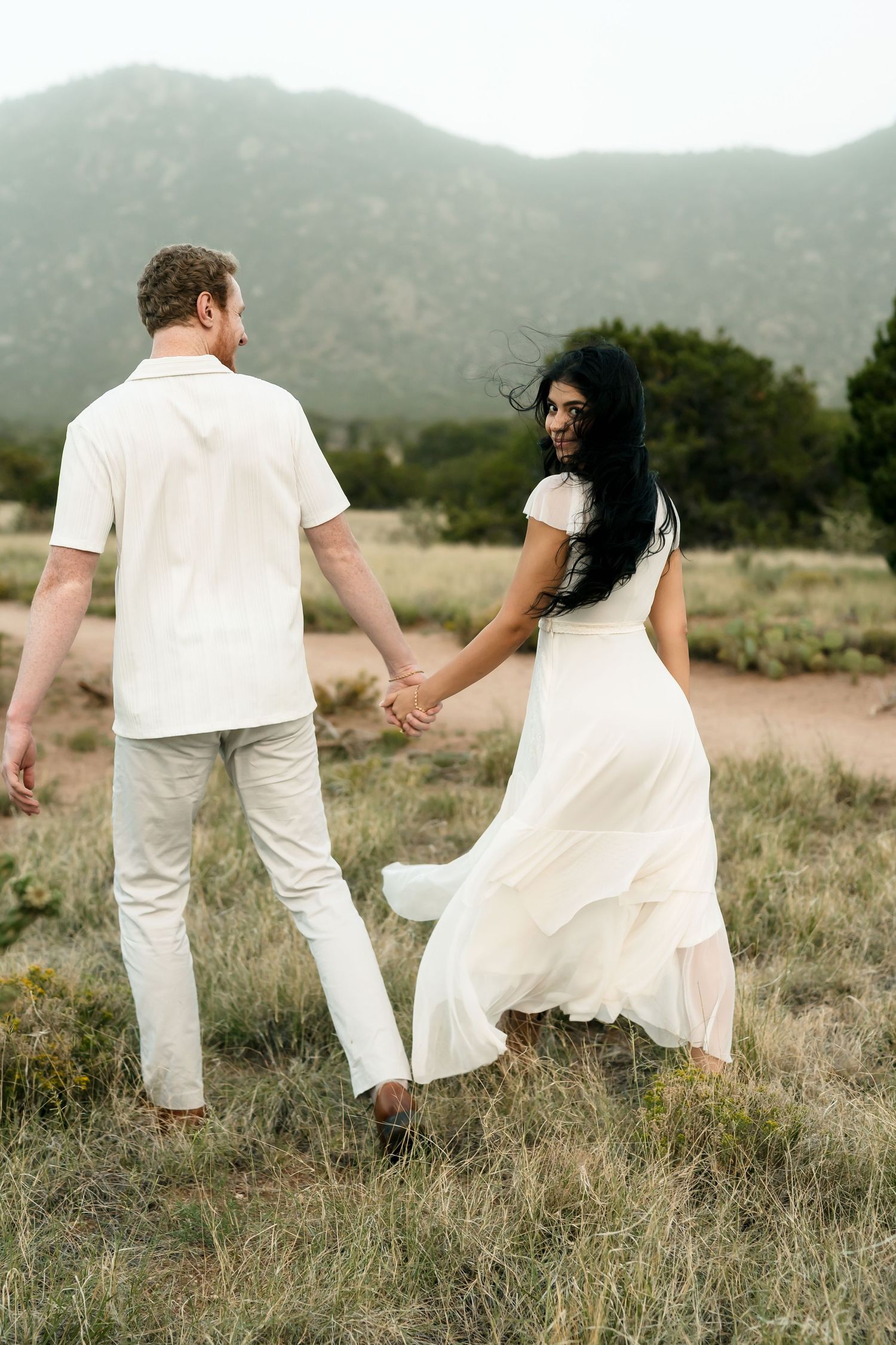 Couple in white outfits holding hands walking through a mountain meadow, woman in flowing dress looking back and smiling.
