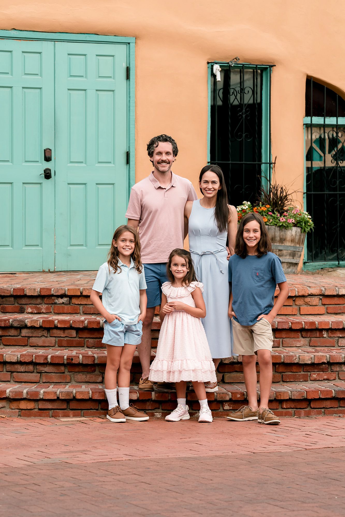 A smiling family of five poses on brick steps in front of a colorful adobe building with turquoise doors.