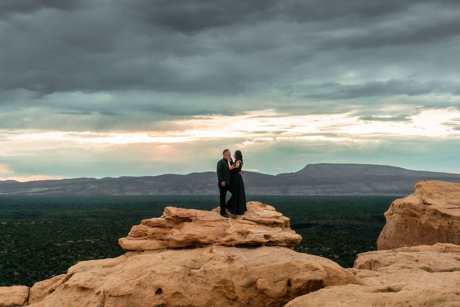 Couple embracing on red rock formation at sunset with dramatic stormy skies and vast desert landscape in background.