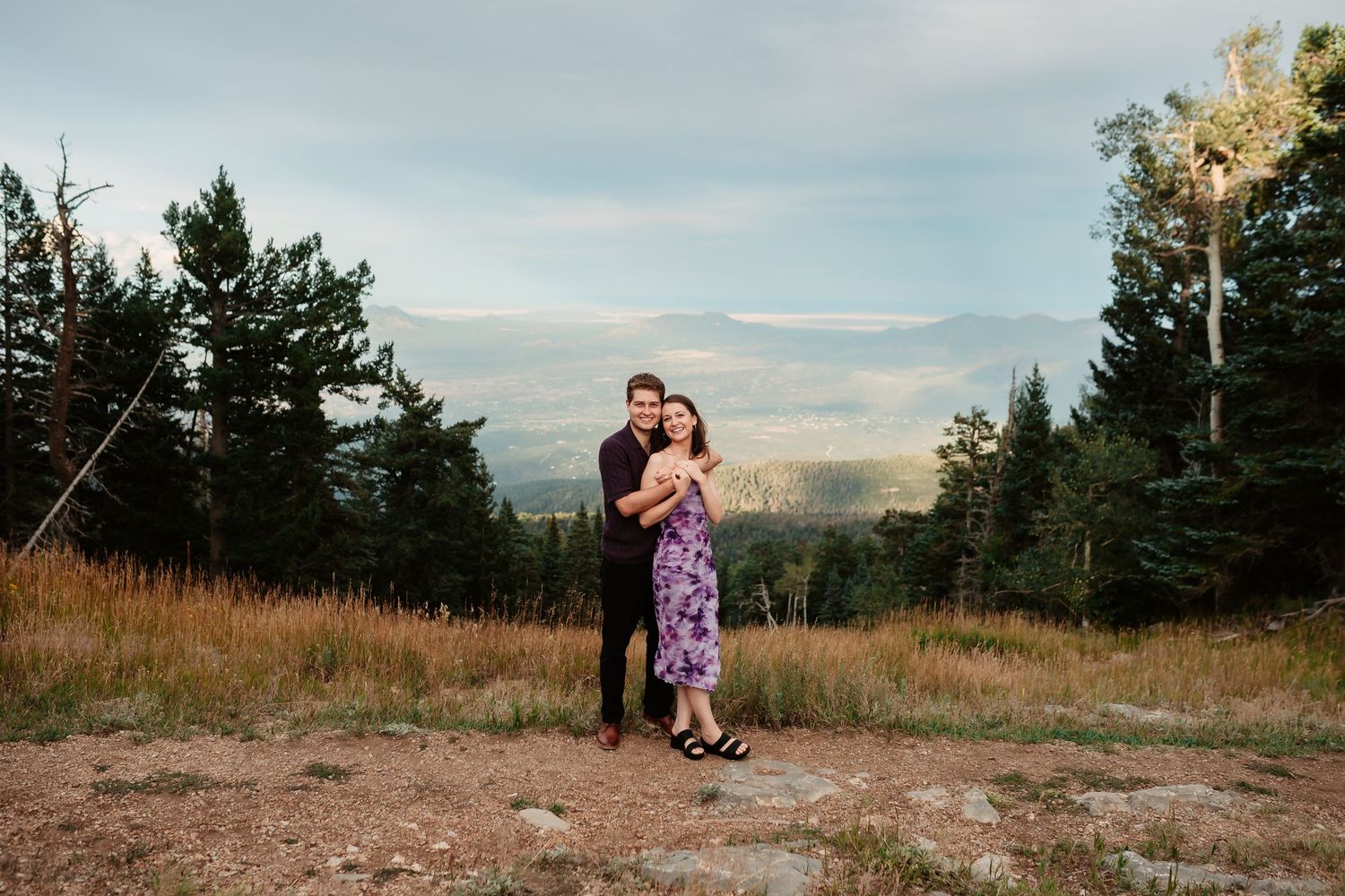 Couple embracing on mountain summit with expansive valley views and pine trees framing the scene.