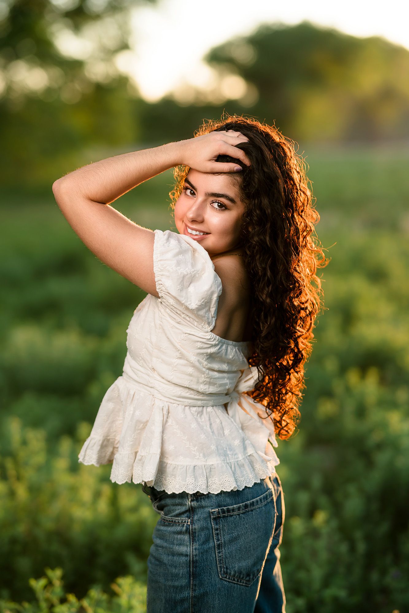 Young woman with curly hair in white top and jeans poses outdoors in green field during golden hour sunset.
