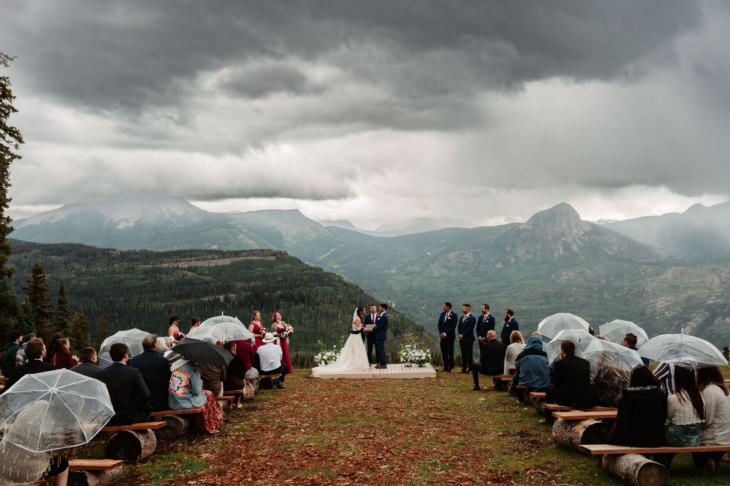 Panoramic view of a mountain wedding ceremony setup with wooden benches against dramatic cloudy skies.