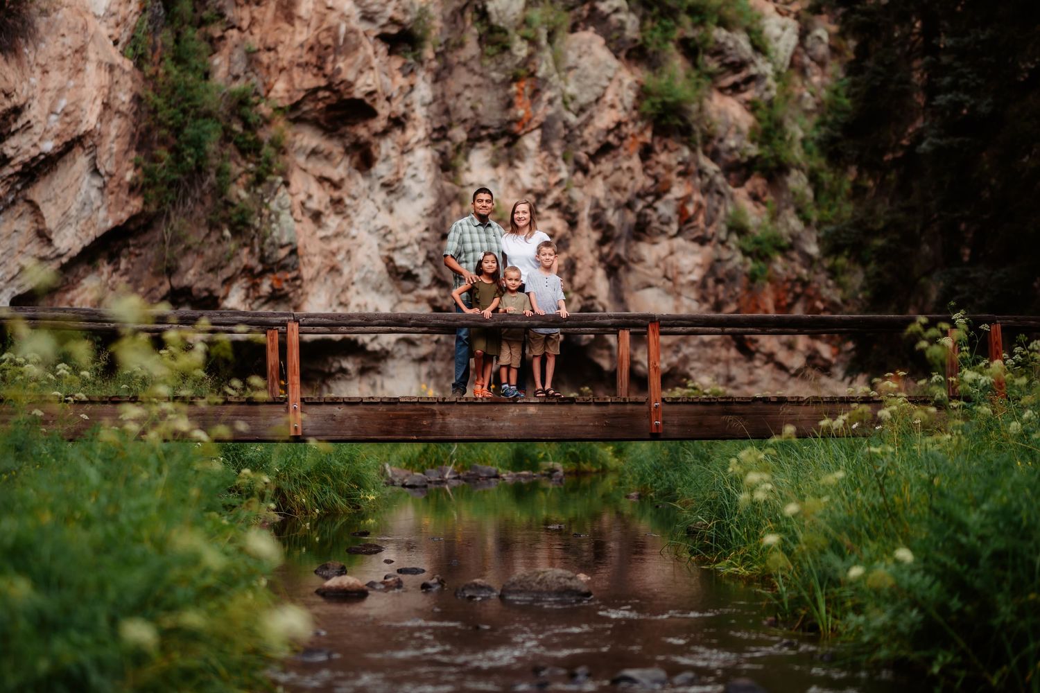 A couple stands on a rustic wooden bridge over a stream with red rock cliffs and green foliage in the background.