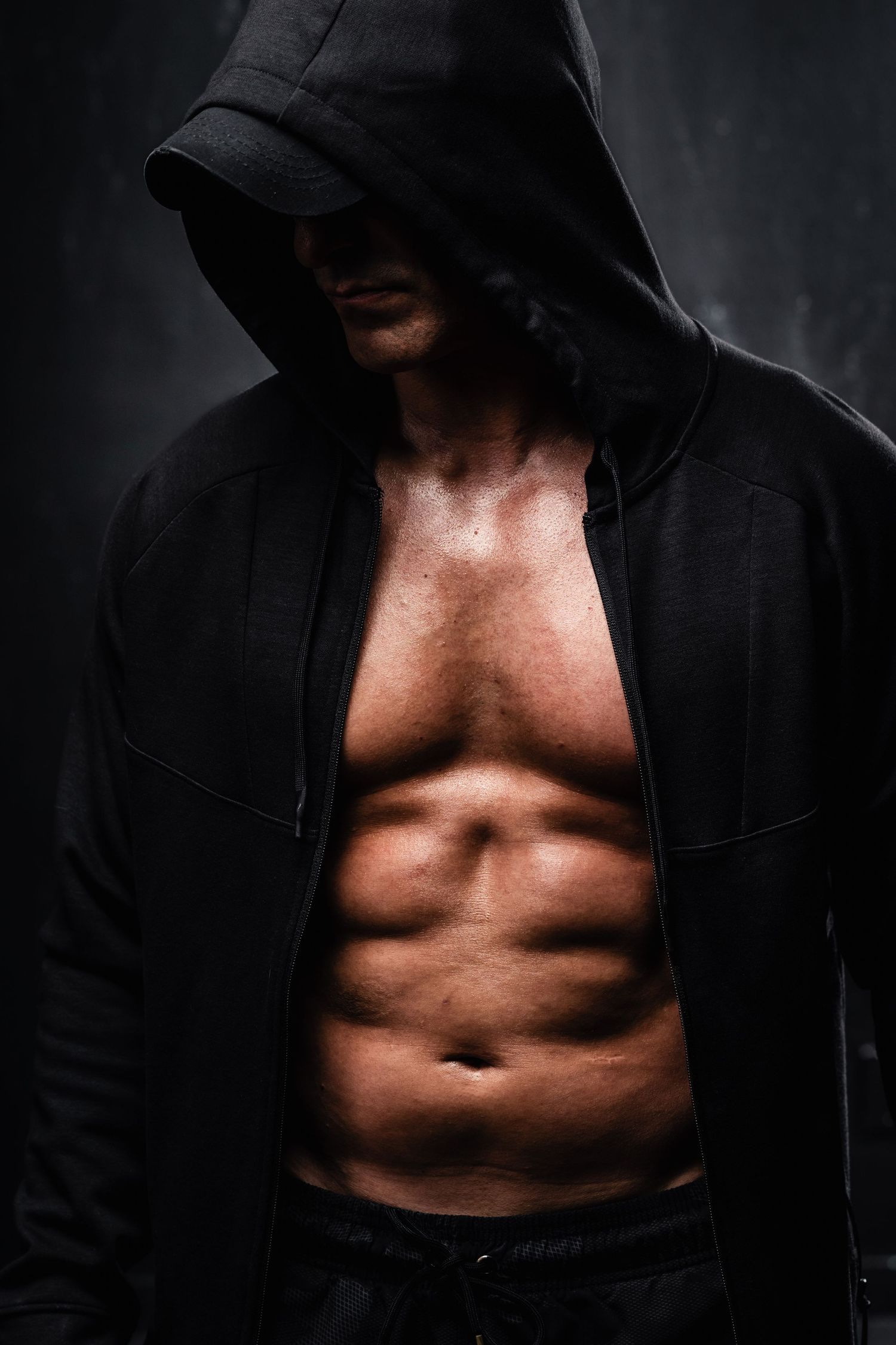 Athletic muscular torso shown in dramatic lighting wearing an open black hoodie against dark background.