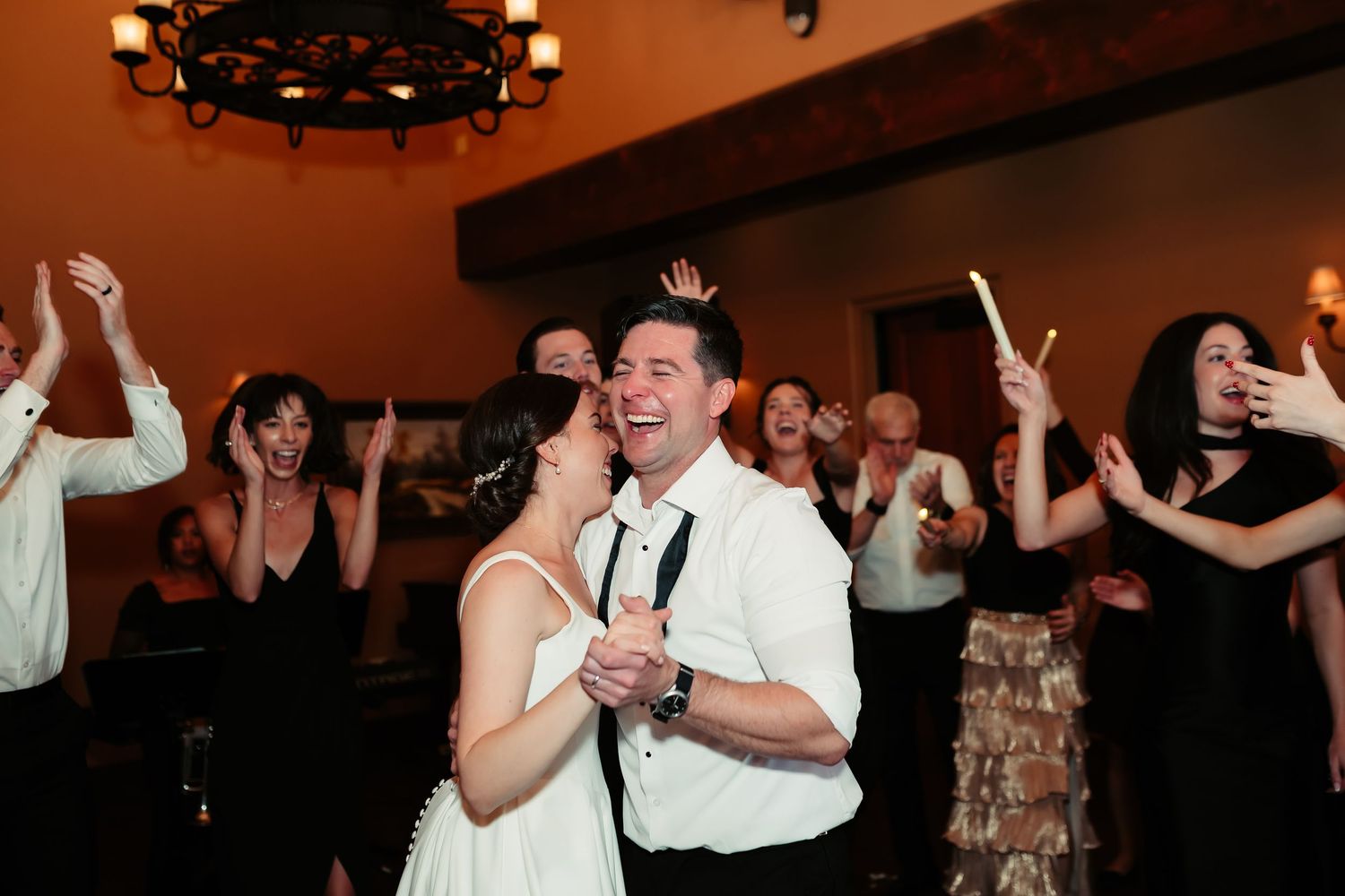 Couples dance together during a wedding reception in a dimly lit ballroom with chandeliers overhead.