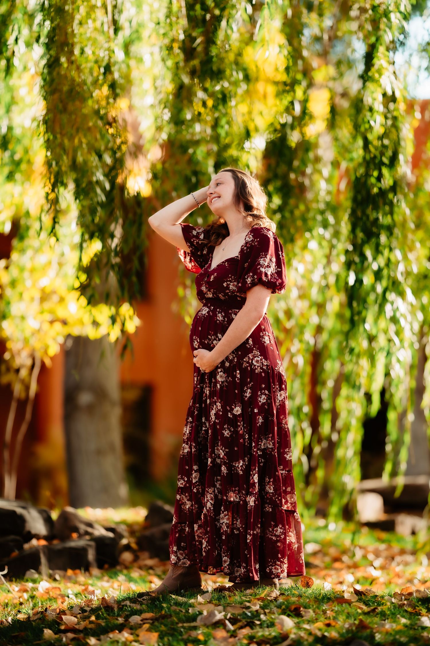 A person in a burgundy velvet dress poses gracefully among autumn leaves and willow trees in a dreamy outdoor setting.