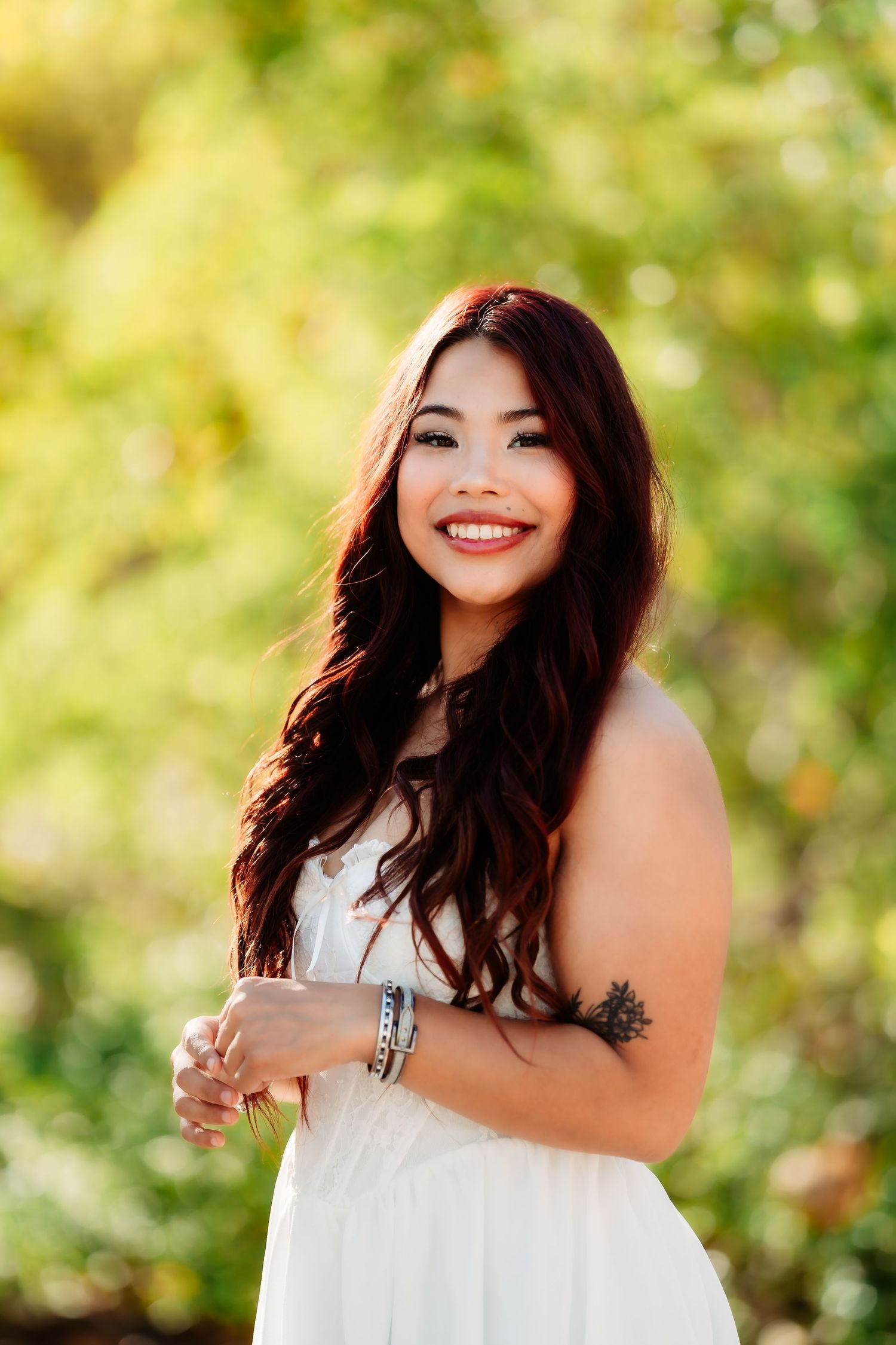 Portrait of graduate in white dress smiling outdoors with natural bokeh background.