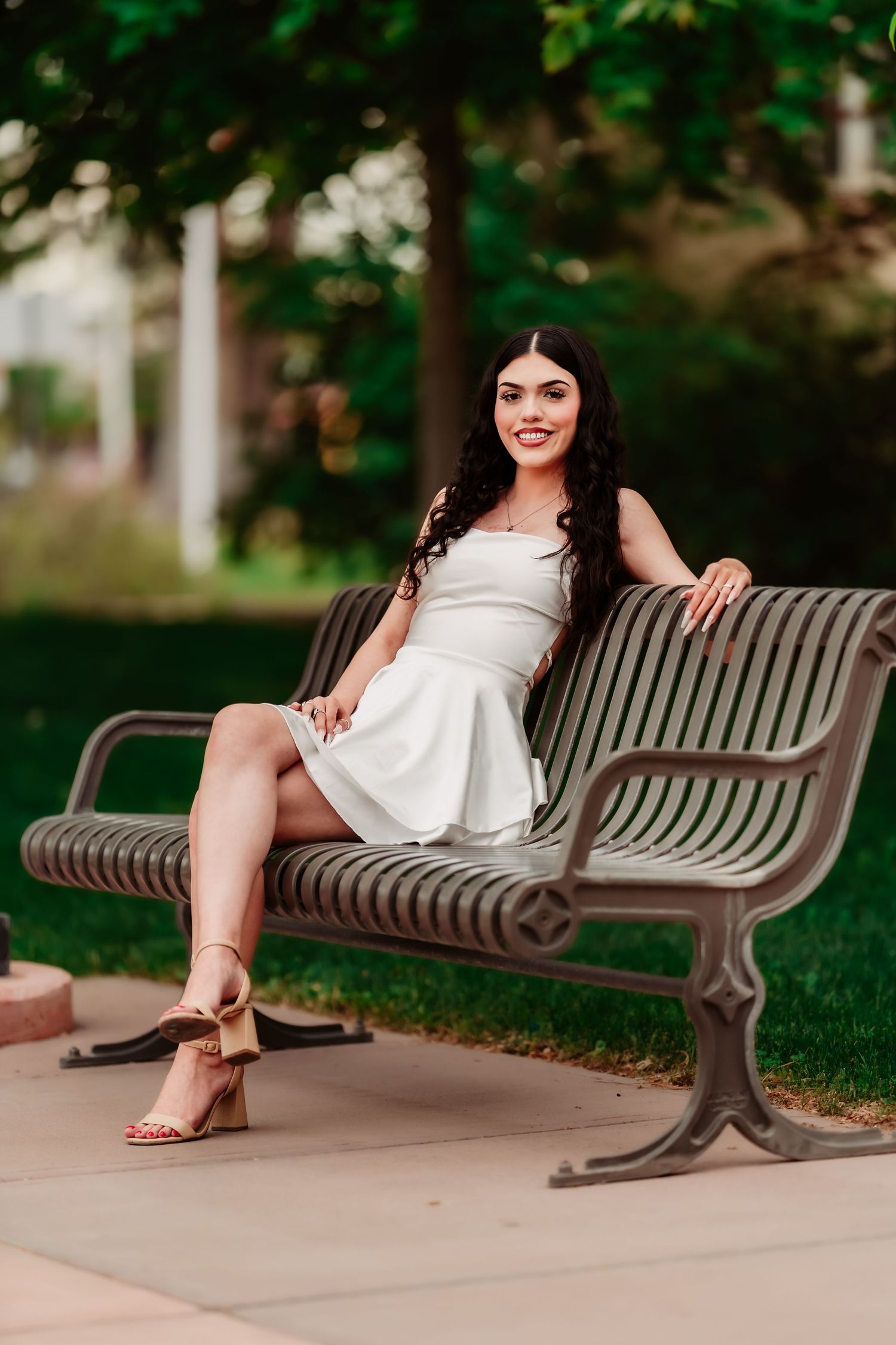 A woman in a white summer dress sits on a metal bench outdoors with trees and greenery in the background.