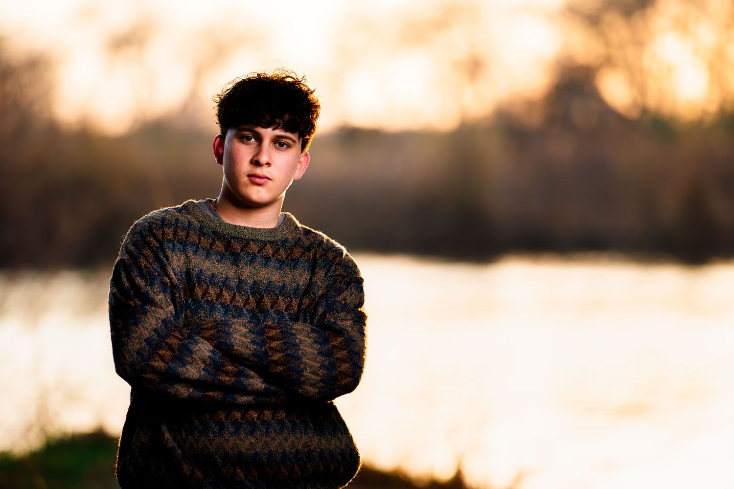 Someone wearing a patterned sweater poses outdoors at golden hour with a blurred natural background.