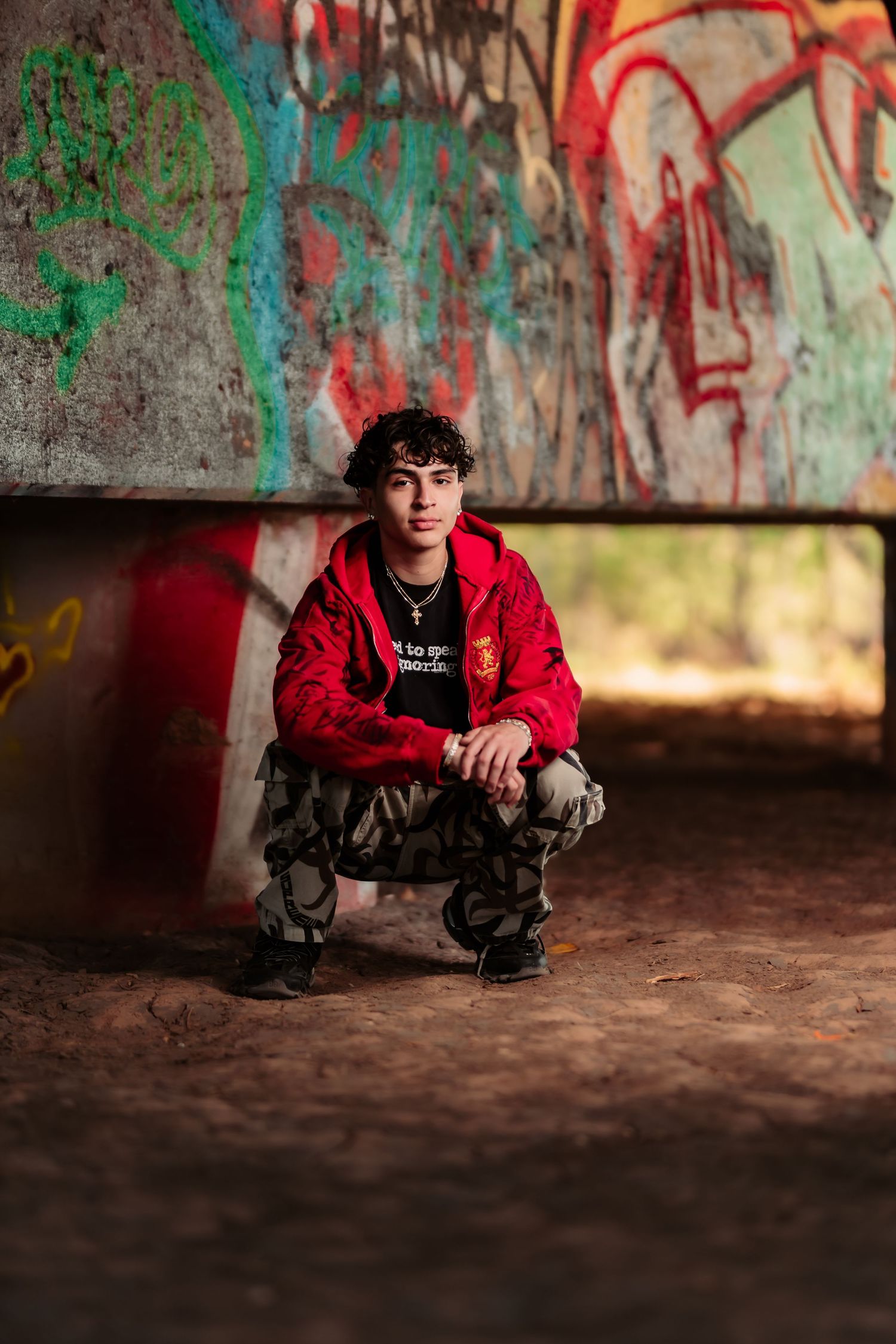 Artistic portrait of someone in a red hoodie crouching against a graffiti wall in an urban setting.