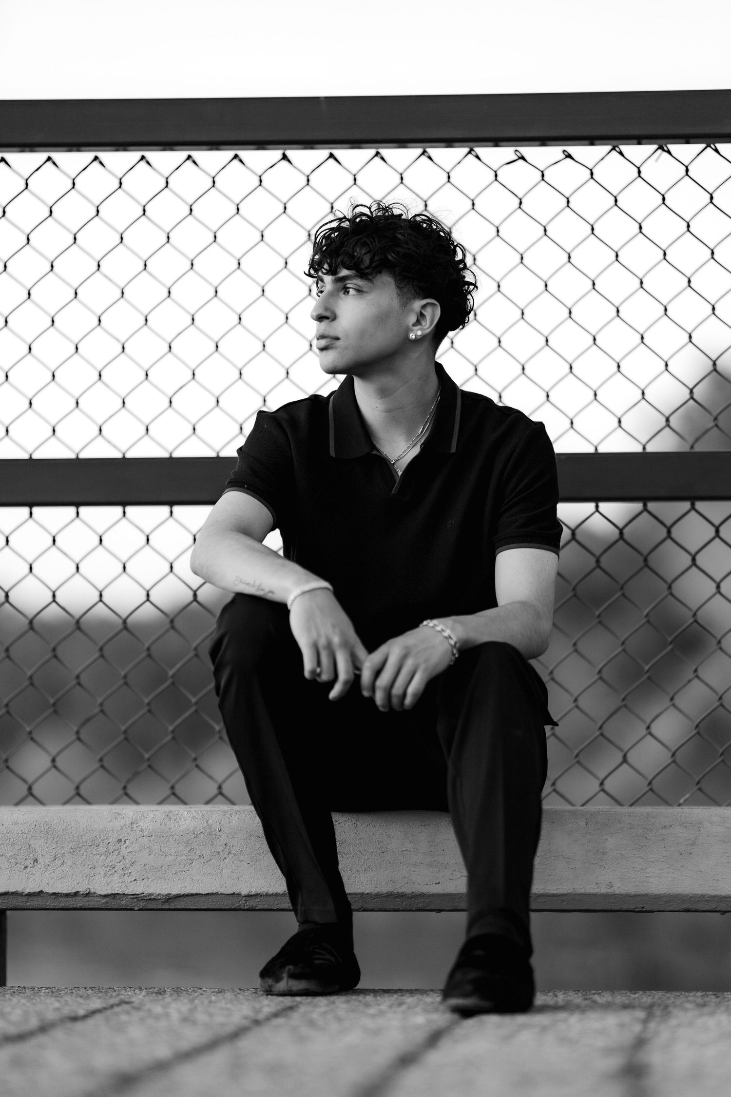 Black and white portrait of a contemplative young person in dark clothing sitting against a chain link fence.