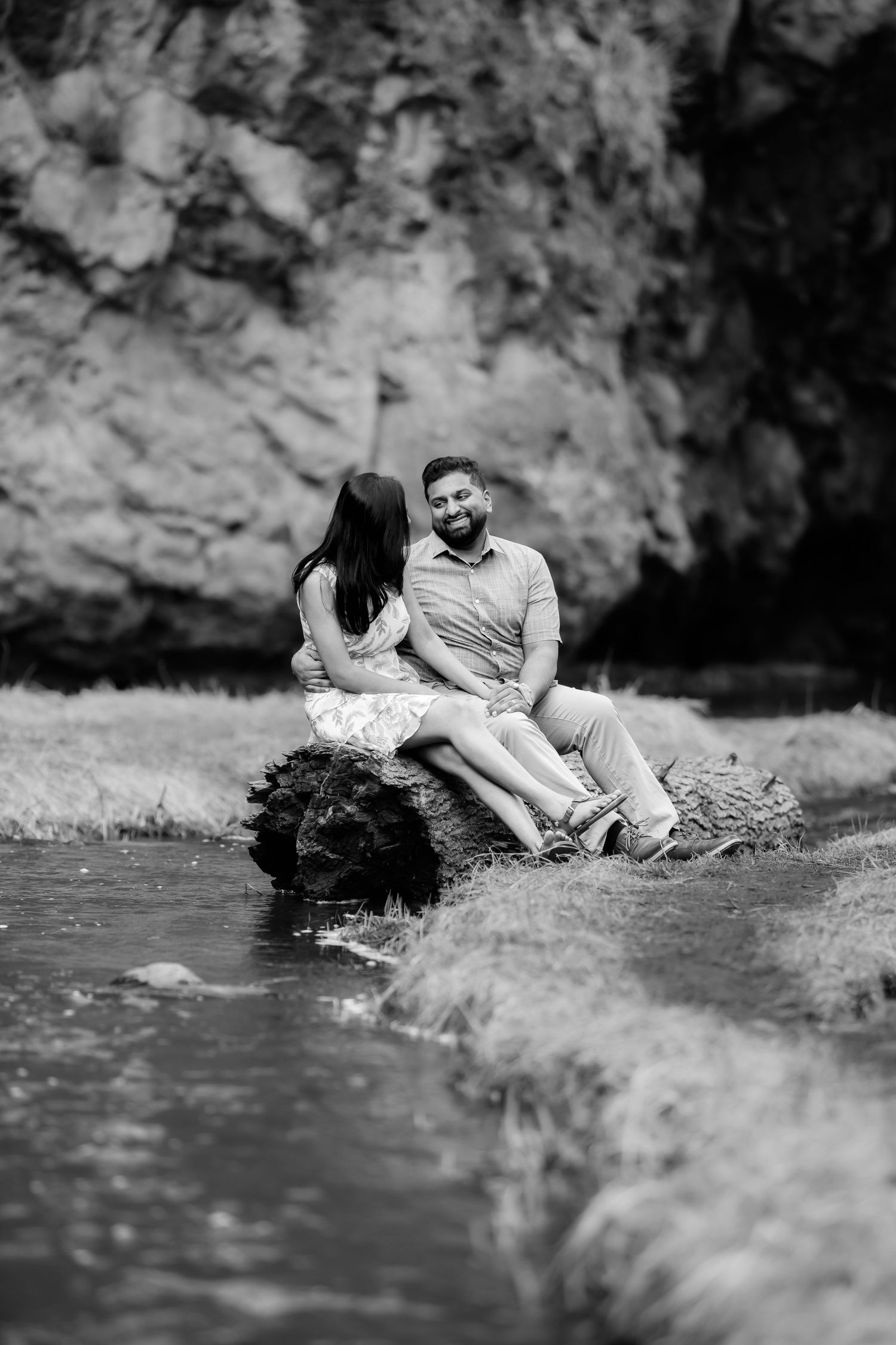 Couple sits together by stream with rocky cliff backdrop in monochrome portrait.