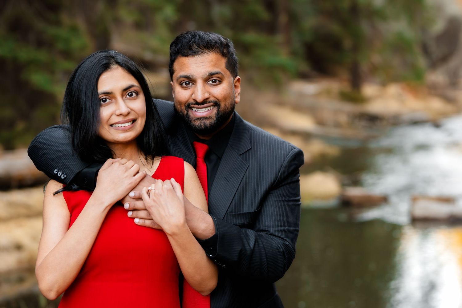 A newly engaged couple smiles while embracing near a serene mountain lake.