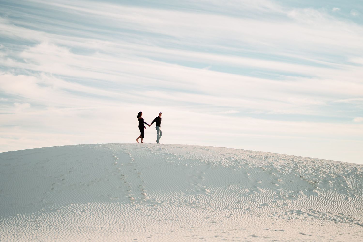 Silhouettes of people walking along a scenic sand dune ridge against a cloudy sky at sunset.