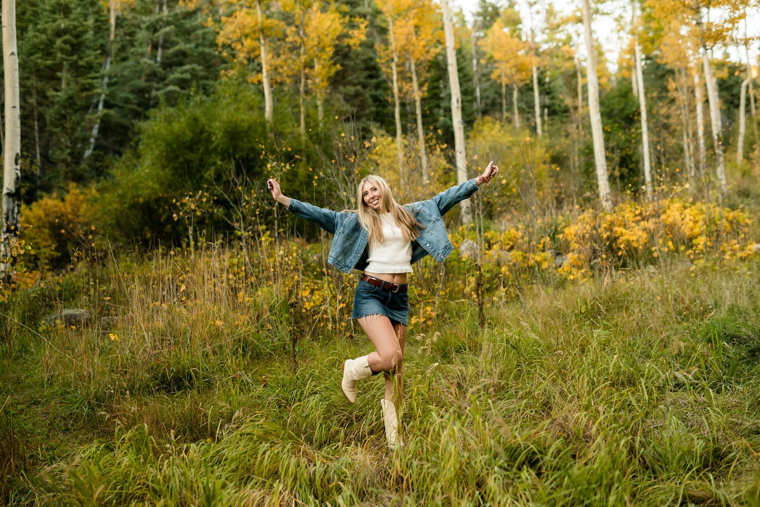 Blonde woman joyfully posing in autumn forest clearing wearing denim jacket, shorts, and boots.