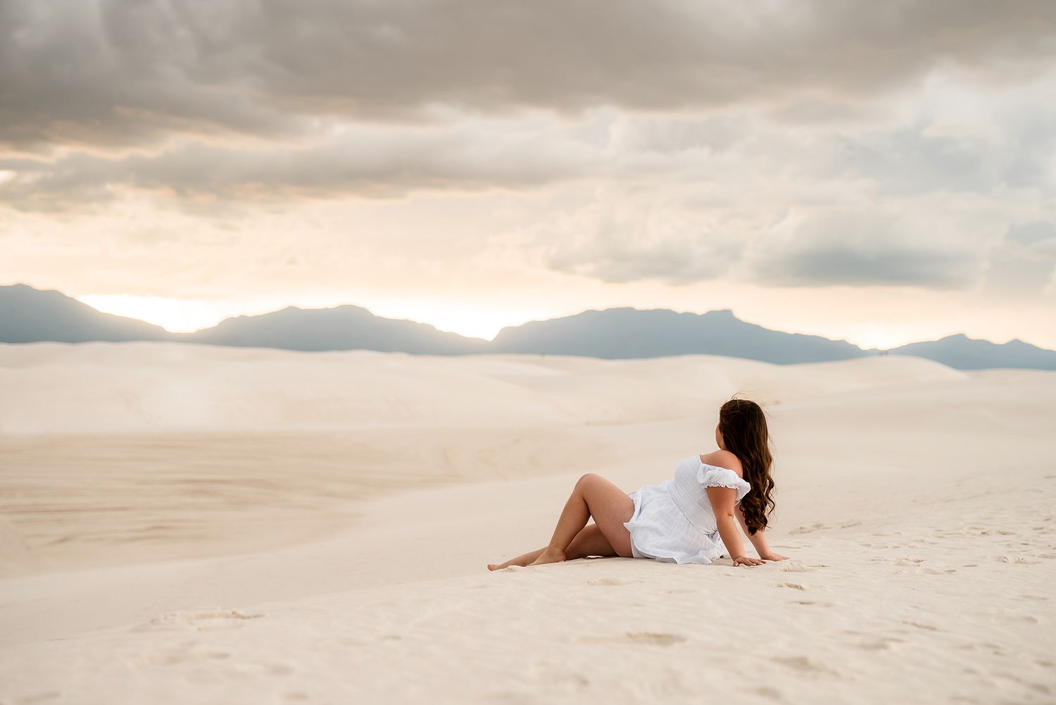 Woman in a white off-the-shoulder dress sitting on pale desert sand dunes at sunset, with distant mountains and soft cloudy skies.