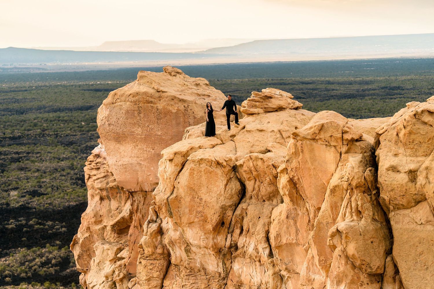 Two figures stand atop a dramatic sandstone rock formation overlooking a vast desert landscape at dusk.