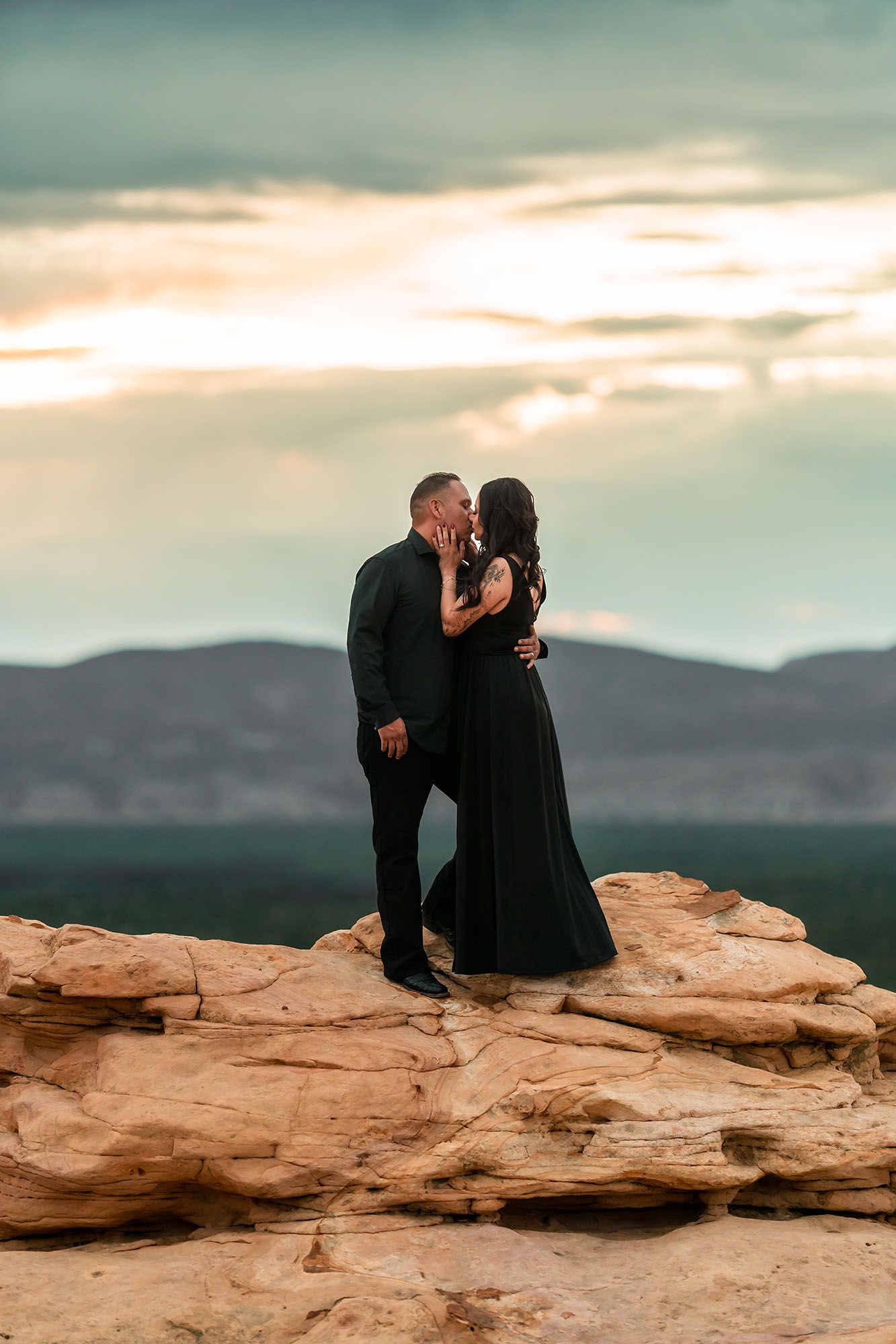 Couple kissing on red rock formation at sunset wearing black formal attire with mountain views.