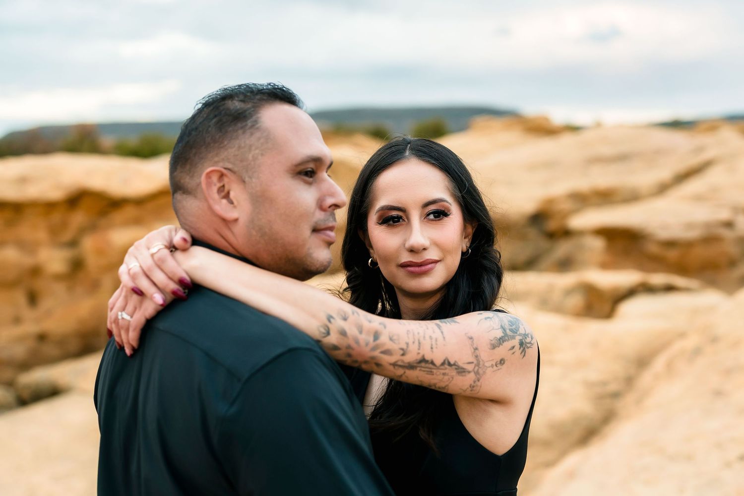 Woman with tattoos embracing man from behind against sandy desert rock backdrop.