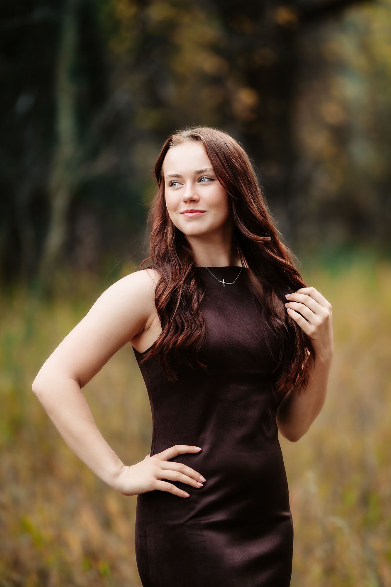 Young woman with long auburn hair in a dark brown sleeveless dress posing outdoors in an autumn forest setting.