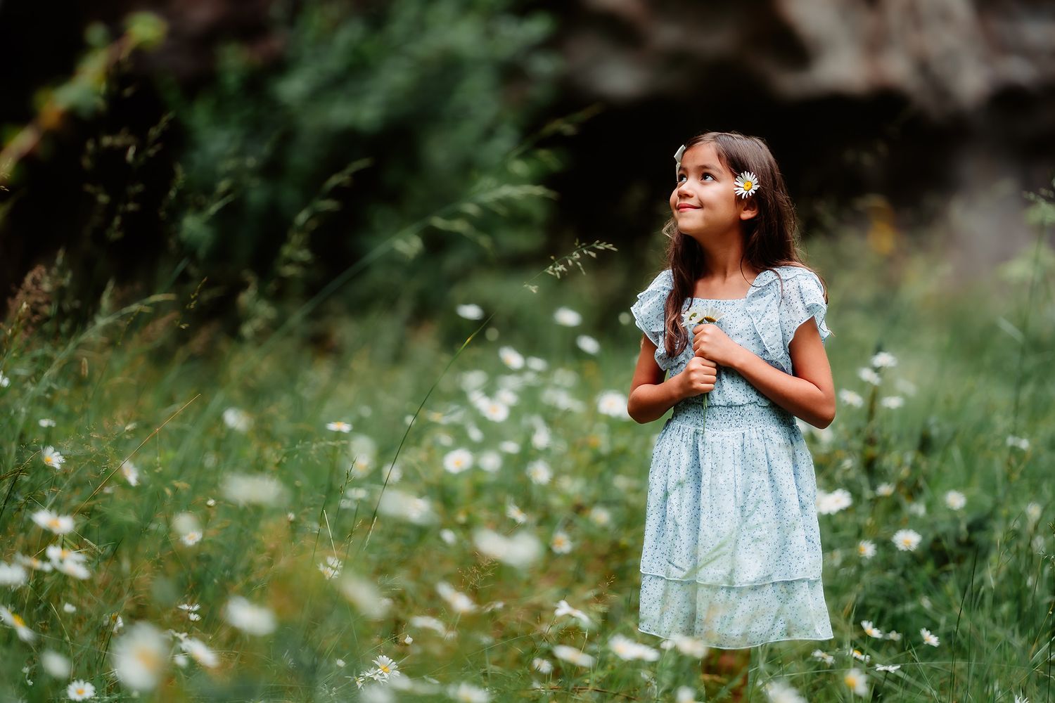 A young girl in a blue floral dress holds wildflowers in a lush green meadow with daisies.