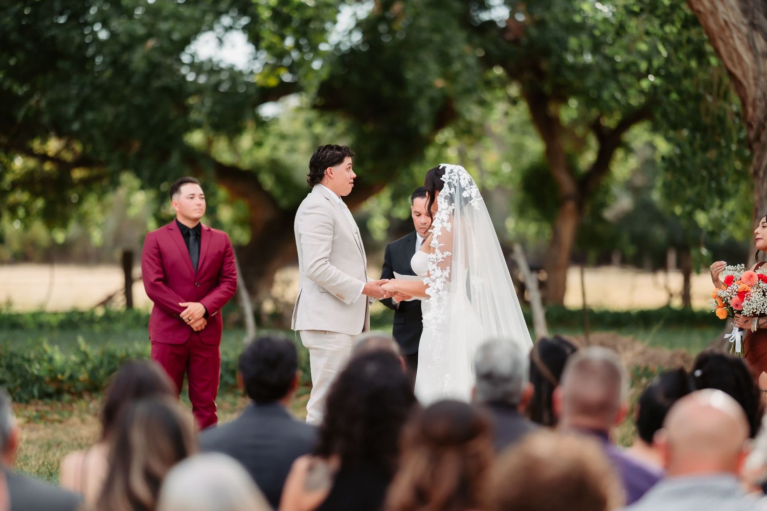 Wedding party gathers outdoors under green trees during summer ceremony celebration.