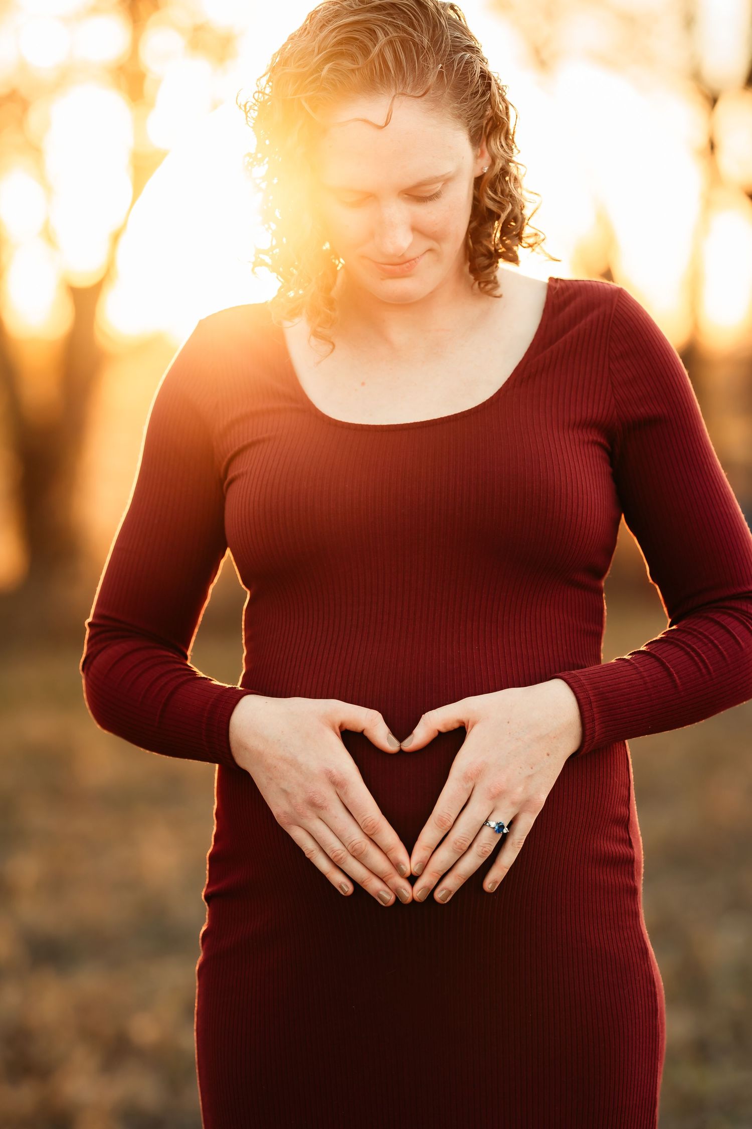A pregnant woman in a burgundy sweater forms a heart shape with her hands over her belly during golden hour.