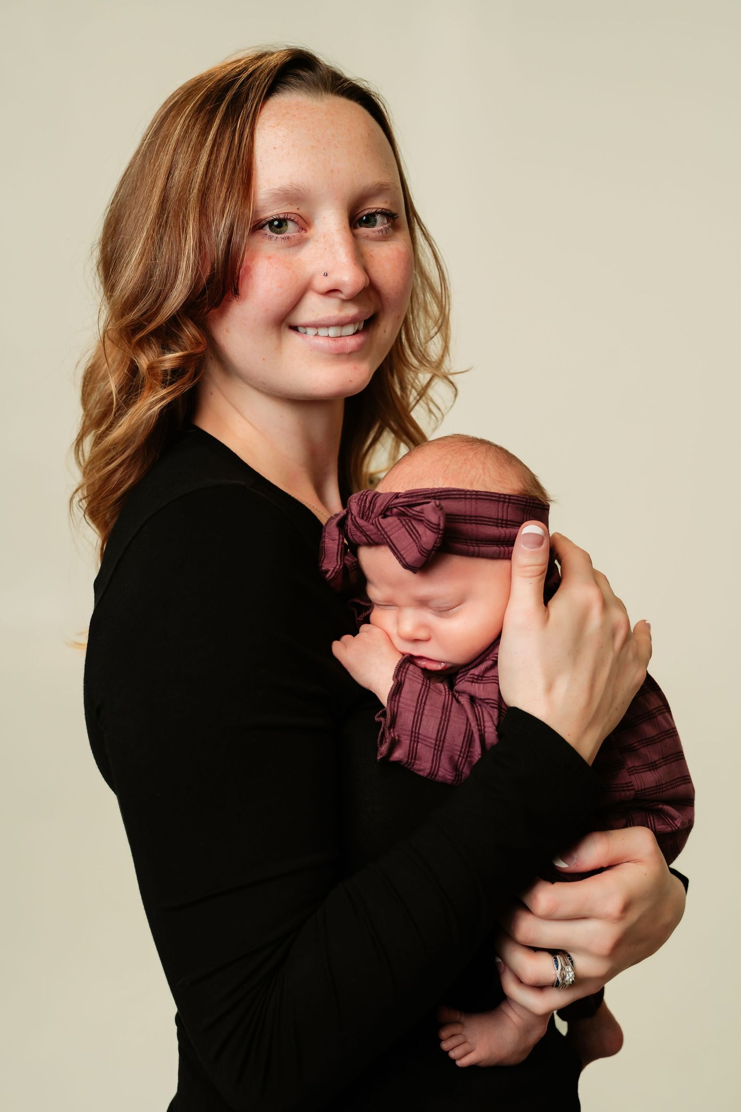A woman in a black sweater holds a sleeping baby wearing a burgundy outfit and matching headband against beige backdrop.