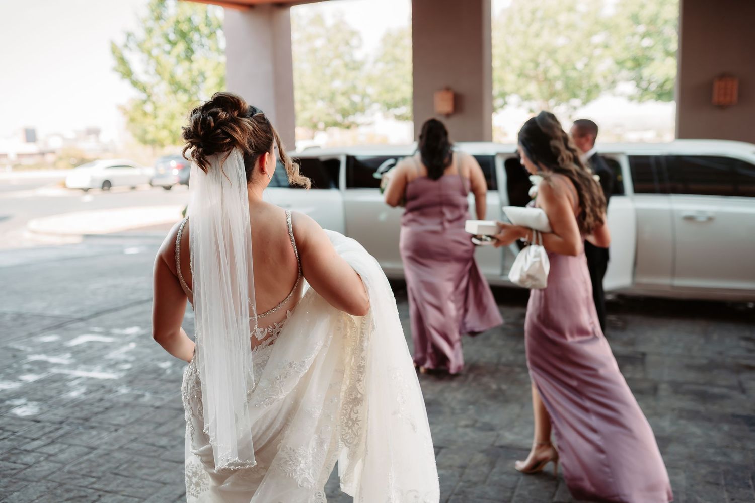 The back of a bride in a flowing white gown walks with bridesmaids in dusty rose dresses outside a venue.