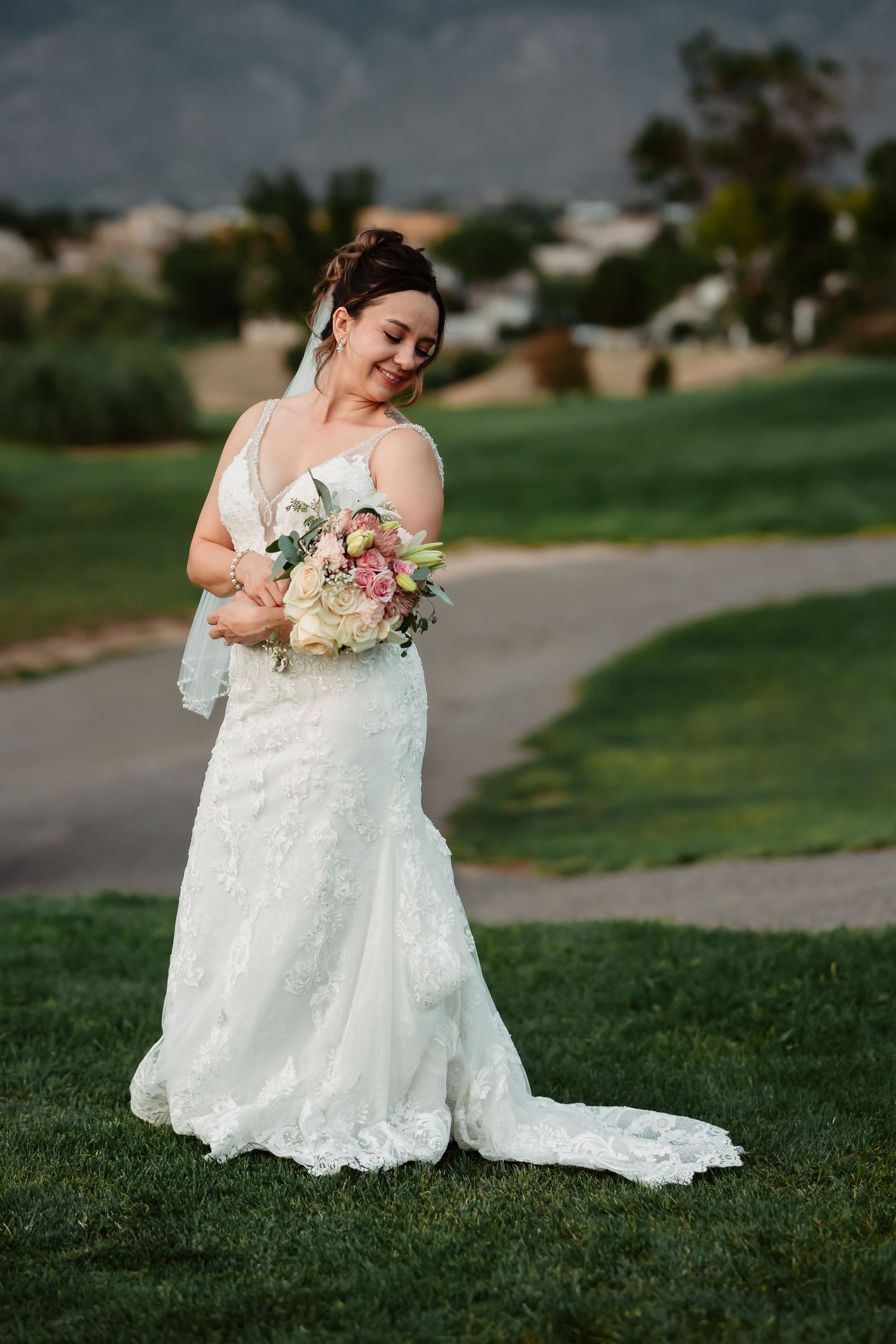 A bride in an elegant white gown sits gracefully on grass with her floral bouquet.