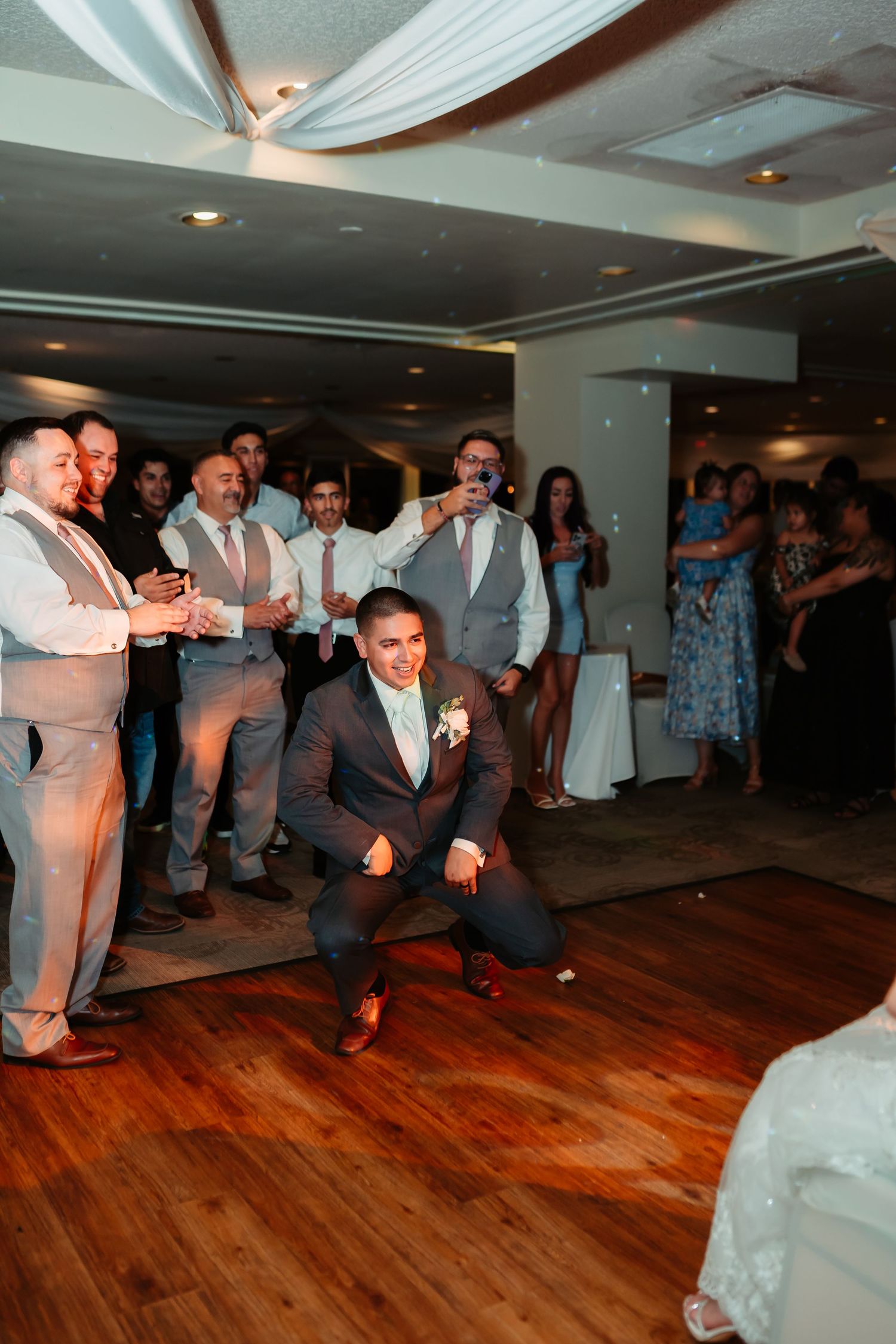 Wedding guests perform a synchronized dance routine on a polished wooden floor under white ceiling draping.