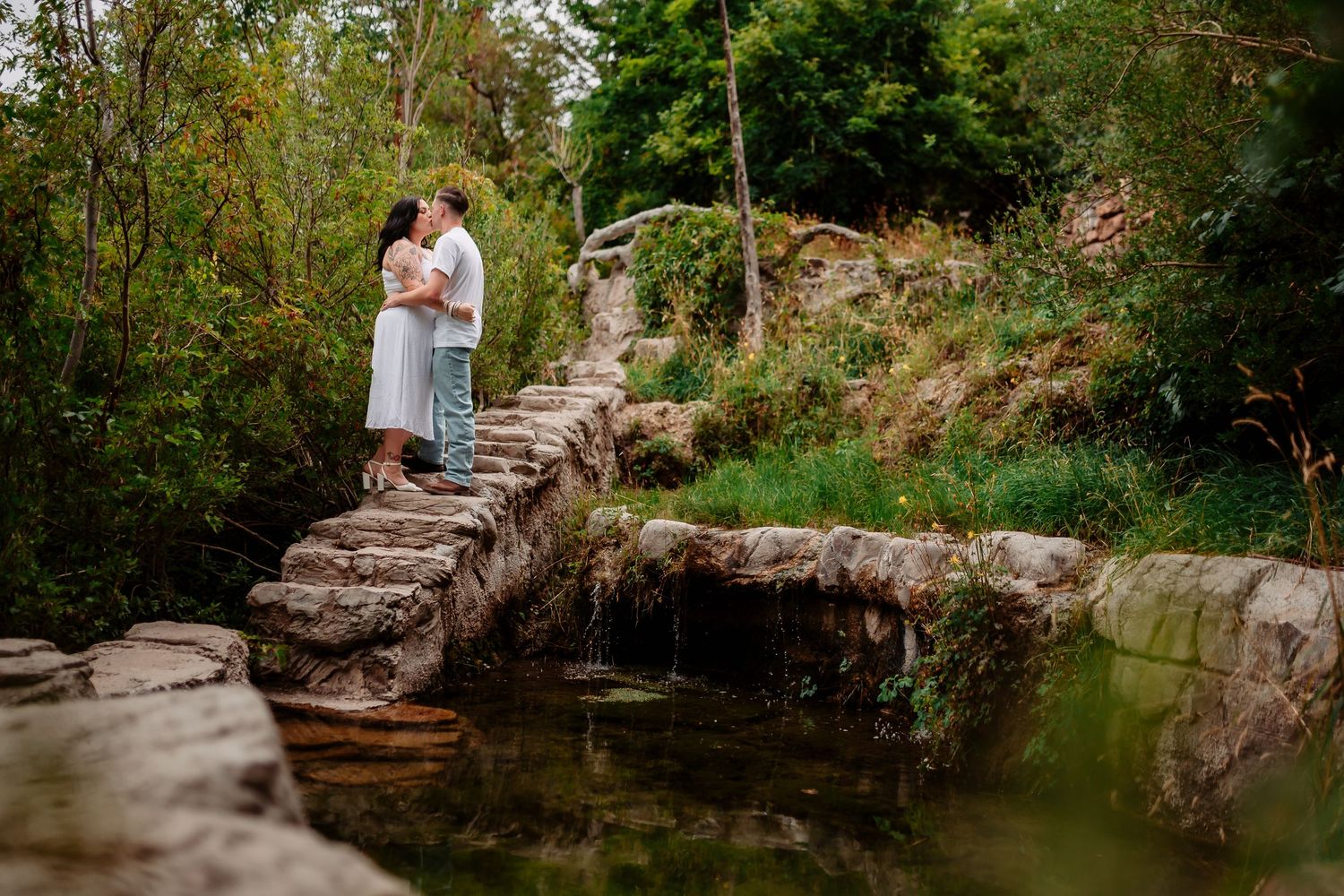 Couple sharing a romantic moment on stone steps by a tranquil pond surrounded by lush greenery.
