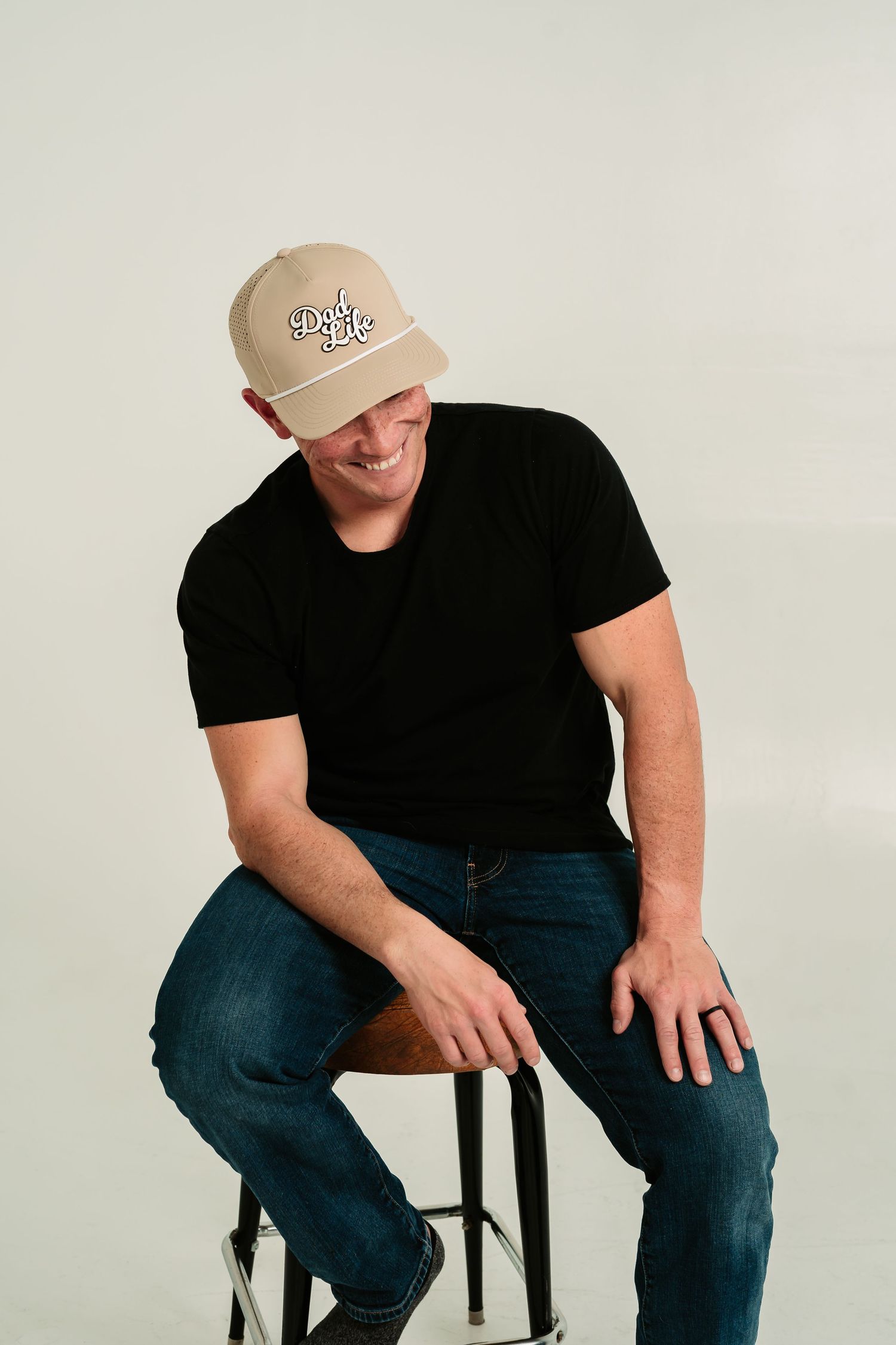 Person wearing baseball cap and black t-shirt sits casually on dark stool against white background.