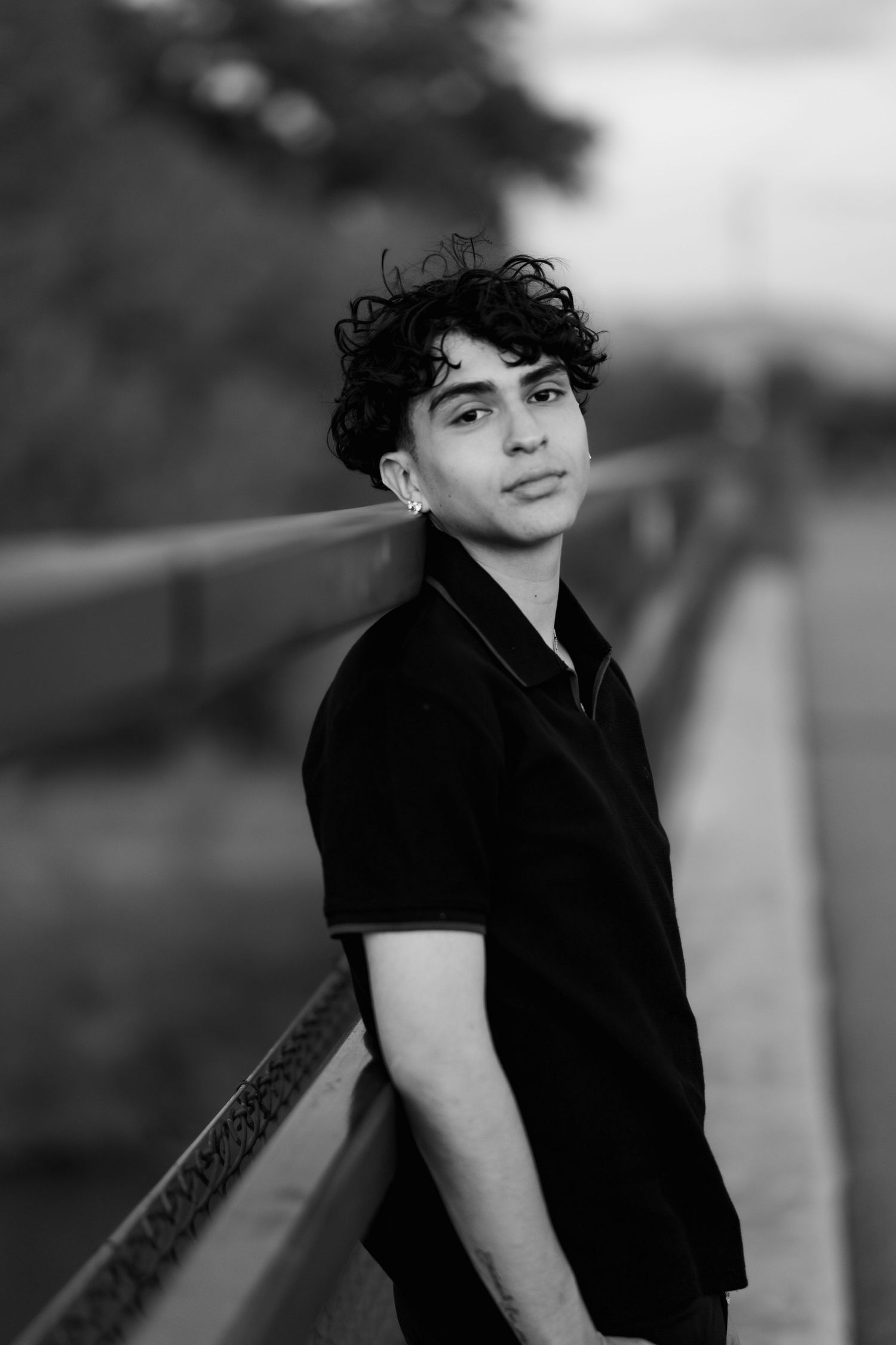 Black and white portrait of a young person with curly dark hair wearing a black polo shirt leaning against a railing outdoors.