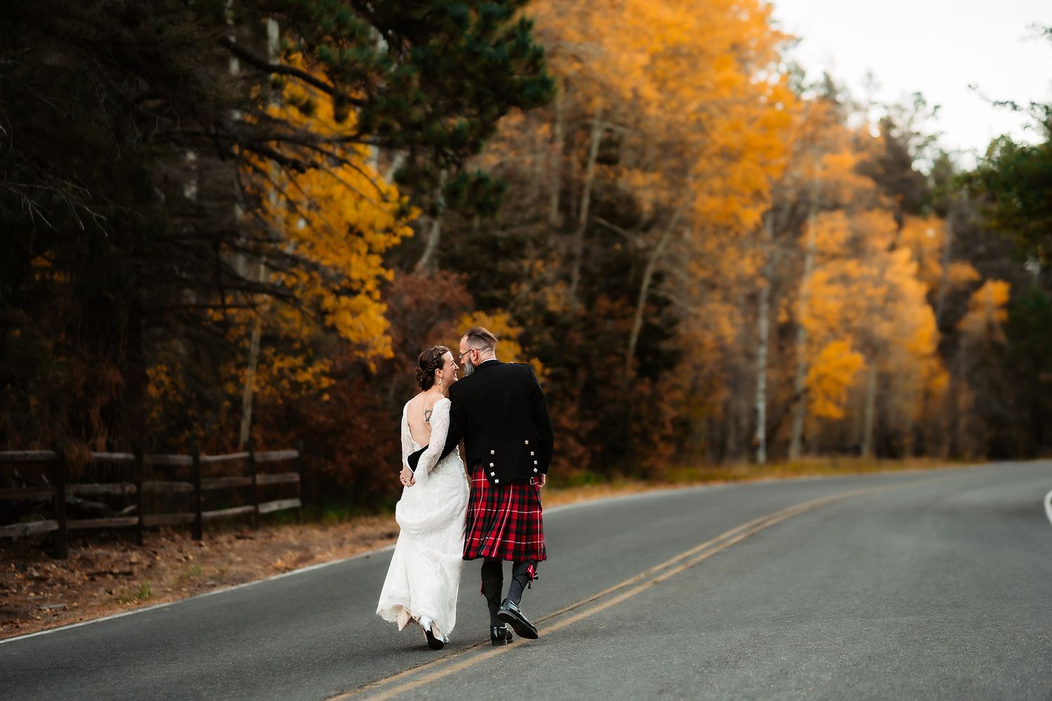 Couple in wedding attire, bride in white gown and groom in red tartan kilt, kissing on a road surrounded by autumn foliage.
