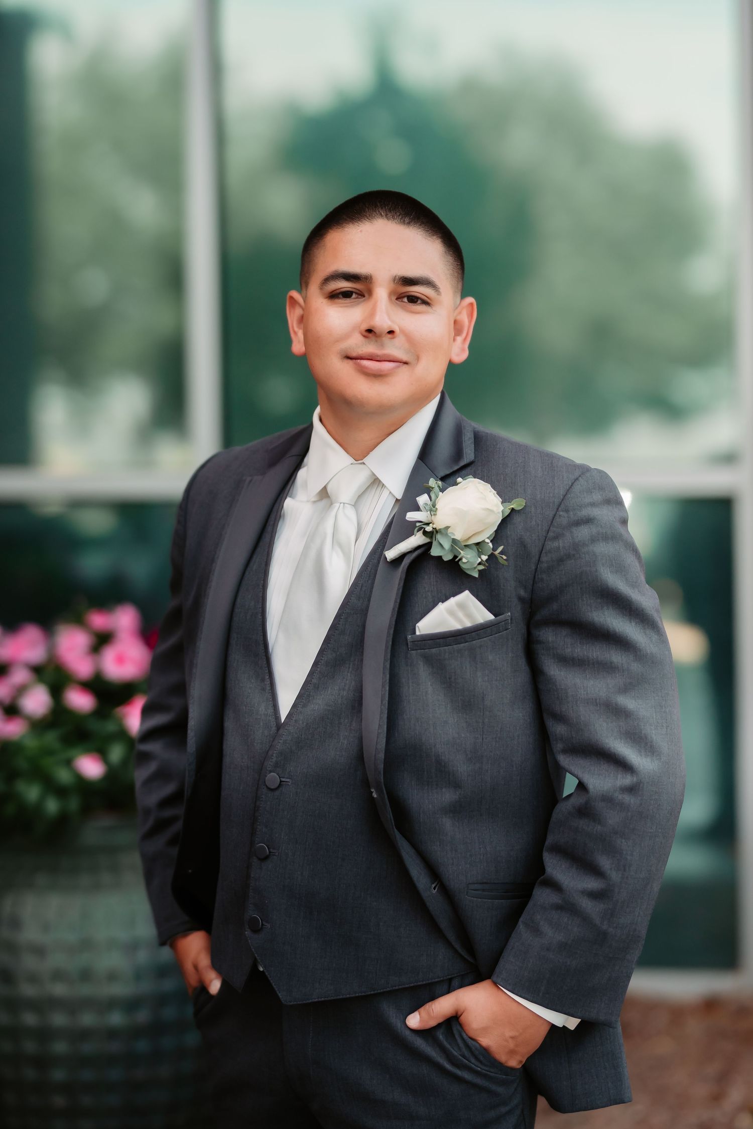 A person in a gray three-piece suit with white boutonniere poses outdoors near pink flowers.