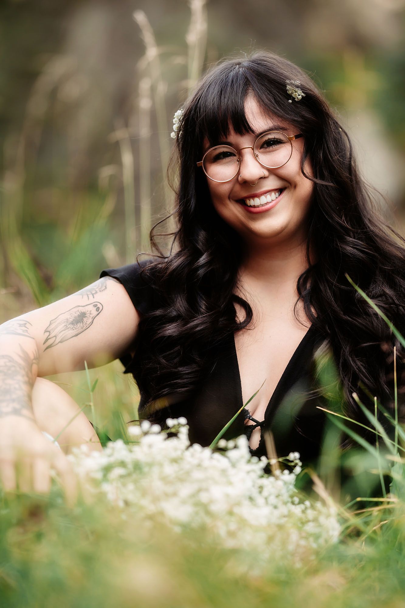 A smiling woman with long dark hair, glasses, and a tattoo sits among wildflowers in a field.