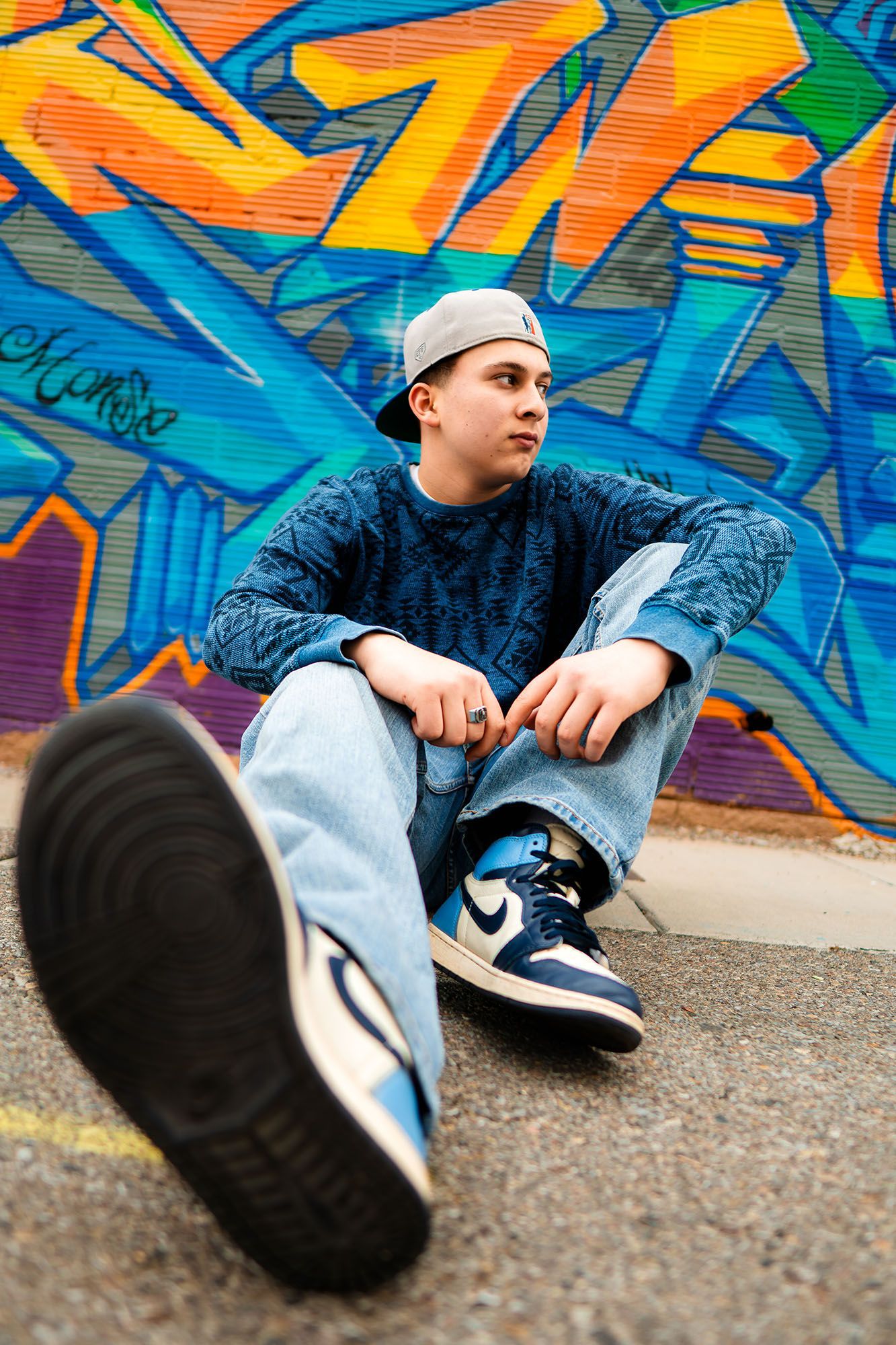 Teen boy in blue sweater, cap, and Nike Air Jordans sitting against colorful graffiti wall.