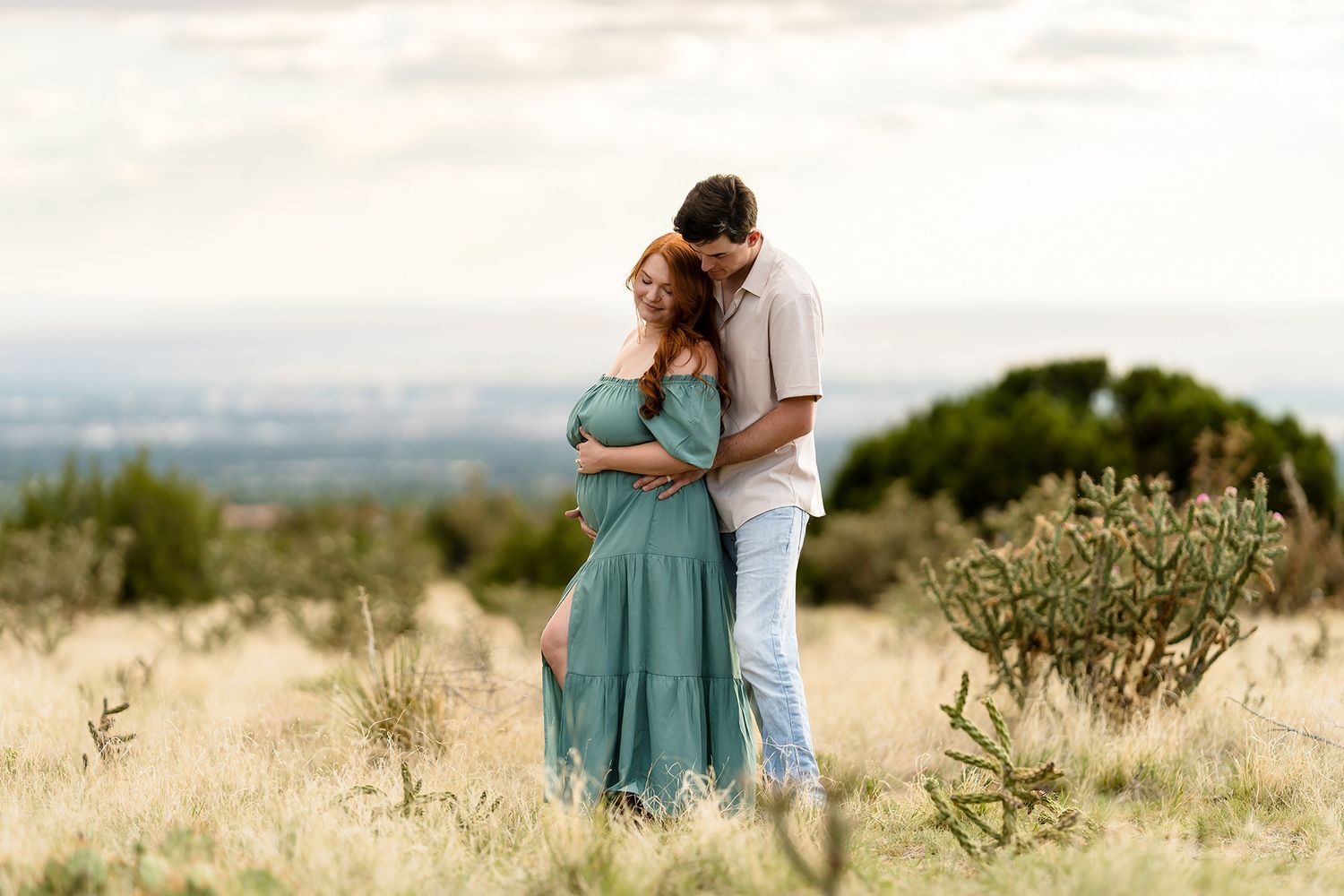 A couple embraces tenderly in a golden field, the woman in a teal dress, with a scenic landscape behind them.